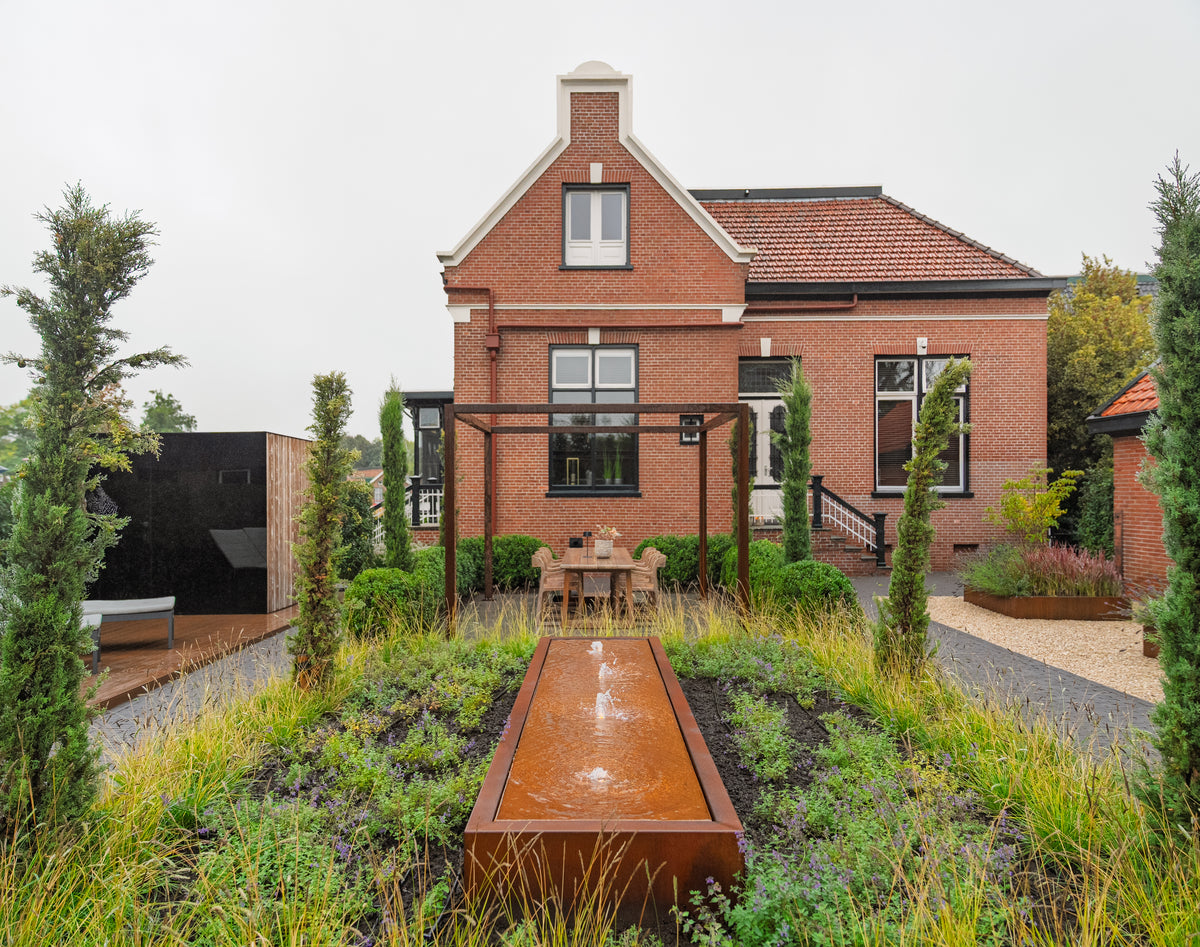 A modern brick house with a landscaped garden featuring a rectangular rusted steel water feature, tall conical plants, and a pergola in the foreground, under an overcast sky.