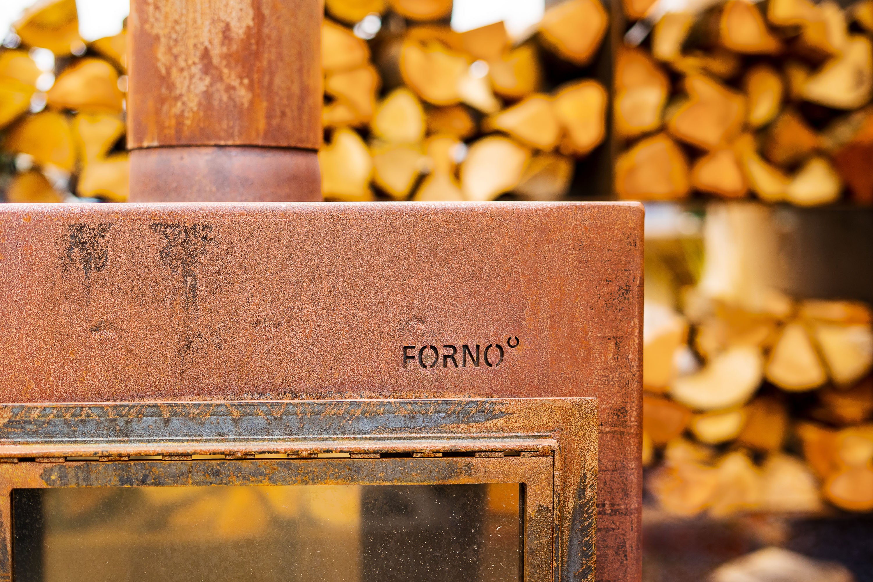 Rust-colored metal object with 'Forno' branding against a background of stacked firewood.