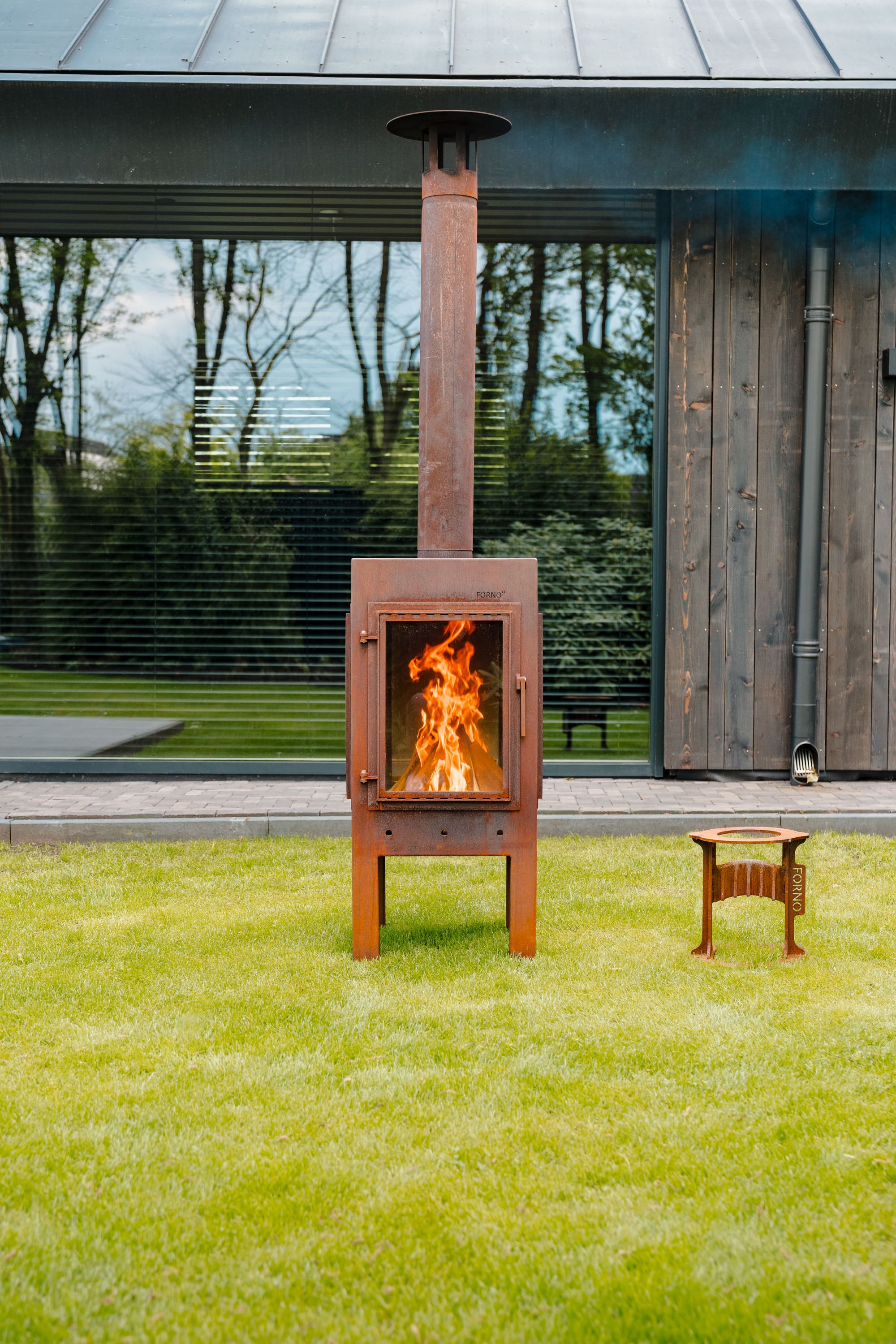 Wood-fired outdoor stove with flames on a grassy area in front of a modern building.