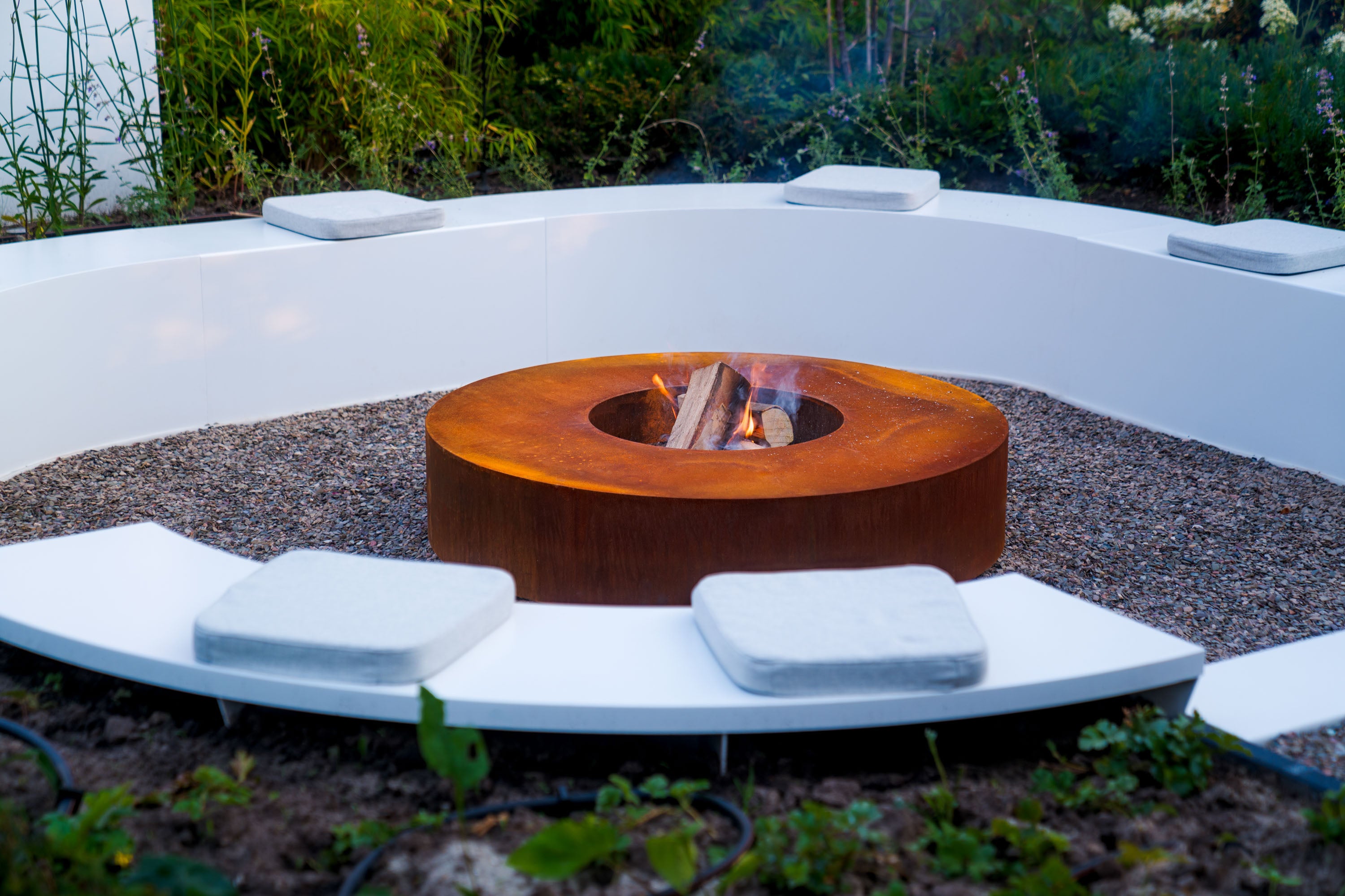 Fire pit in a modern outdoor setting with white seating area and greenery.
