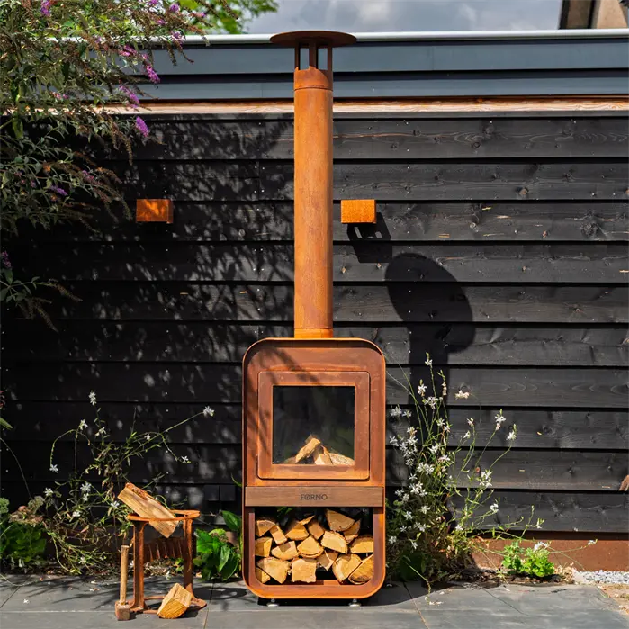 A rusted orange outdoor wood-burning stove with a tall chimney pipe, set against a dark wooden fence with some plants and stacked firewood visible.