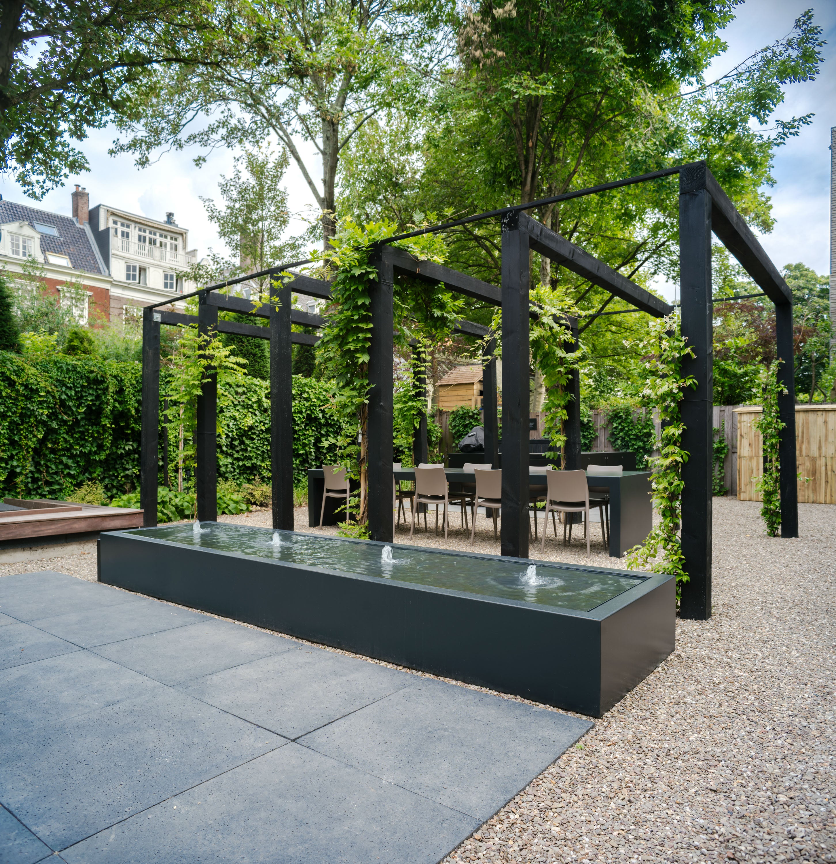 black rectangle water table with four fountains filled with water next to a pergola with seating