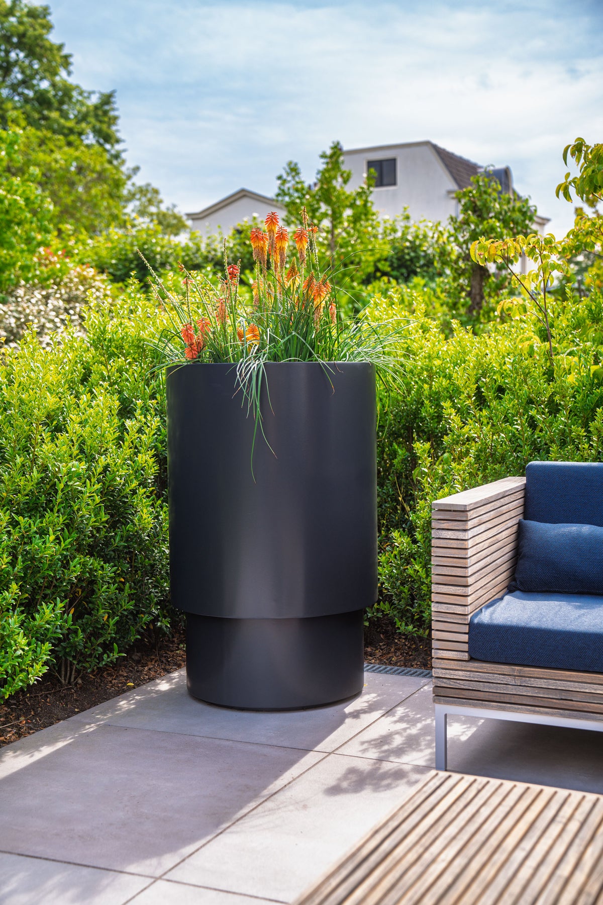A modern black cylindrical planter with layered tiers, filled with green grass and orange flowers, set on a stone patio in a lush garden next to a wooden outdoor sofa with blue cushions.