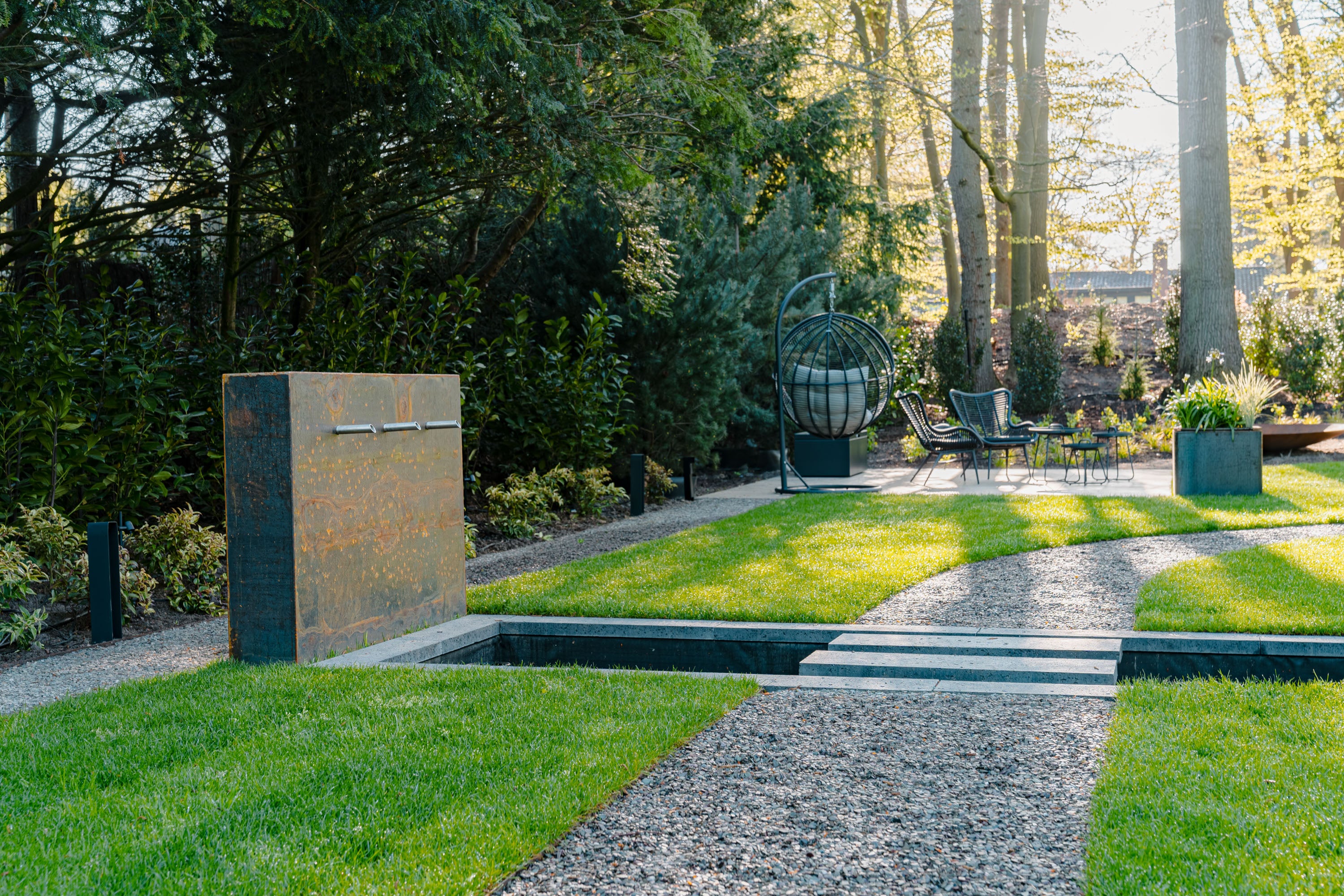 A modern outdoor garden scene with a gravel path leading through manicured green lawns toward patio furniture, a tall rectangular stone wall feature on the left, and dense trees in the background.