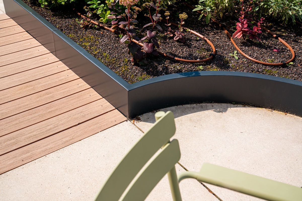 Modern outdoor garden bed with a curved dark gray metal planter border, a light wood deck on the left, a concrete patio in the foreground, and a green chair partially visible at the bottom right.