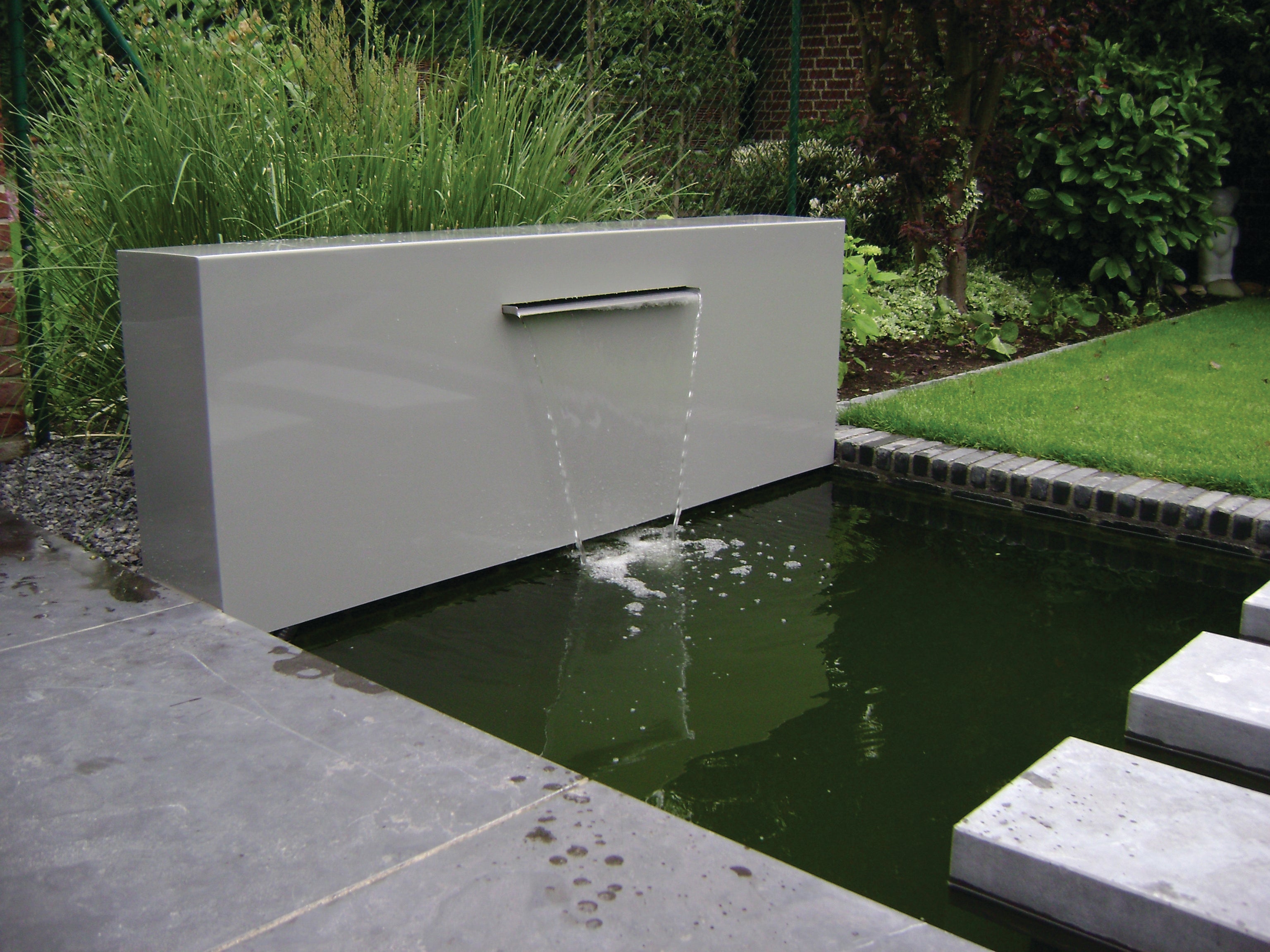 A modern rectangular outdoor wall fountain with water cascading into a small pond, set in a landscaped garden with grass, plants, and stone paving.