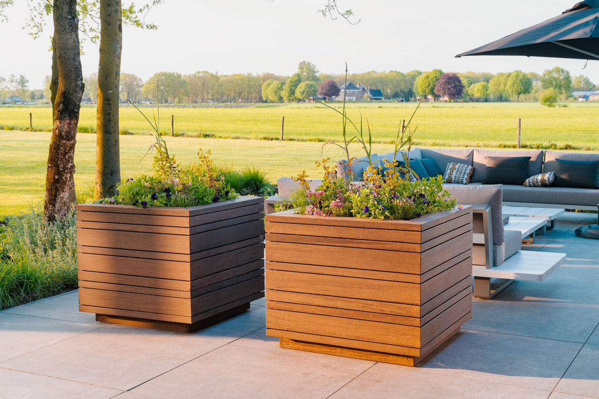 Two large square wooden planter boxes filled with green and yellow plants, placed on a stone patio next to outdoor patio furniture with a grassy field in the background.