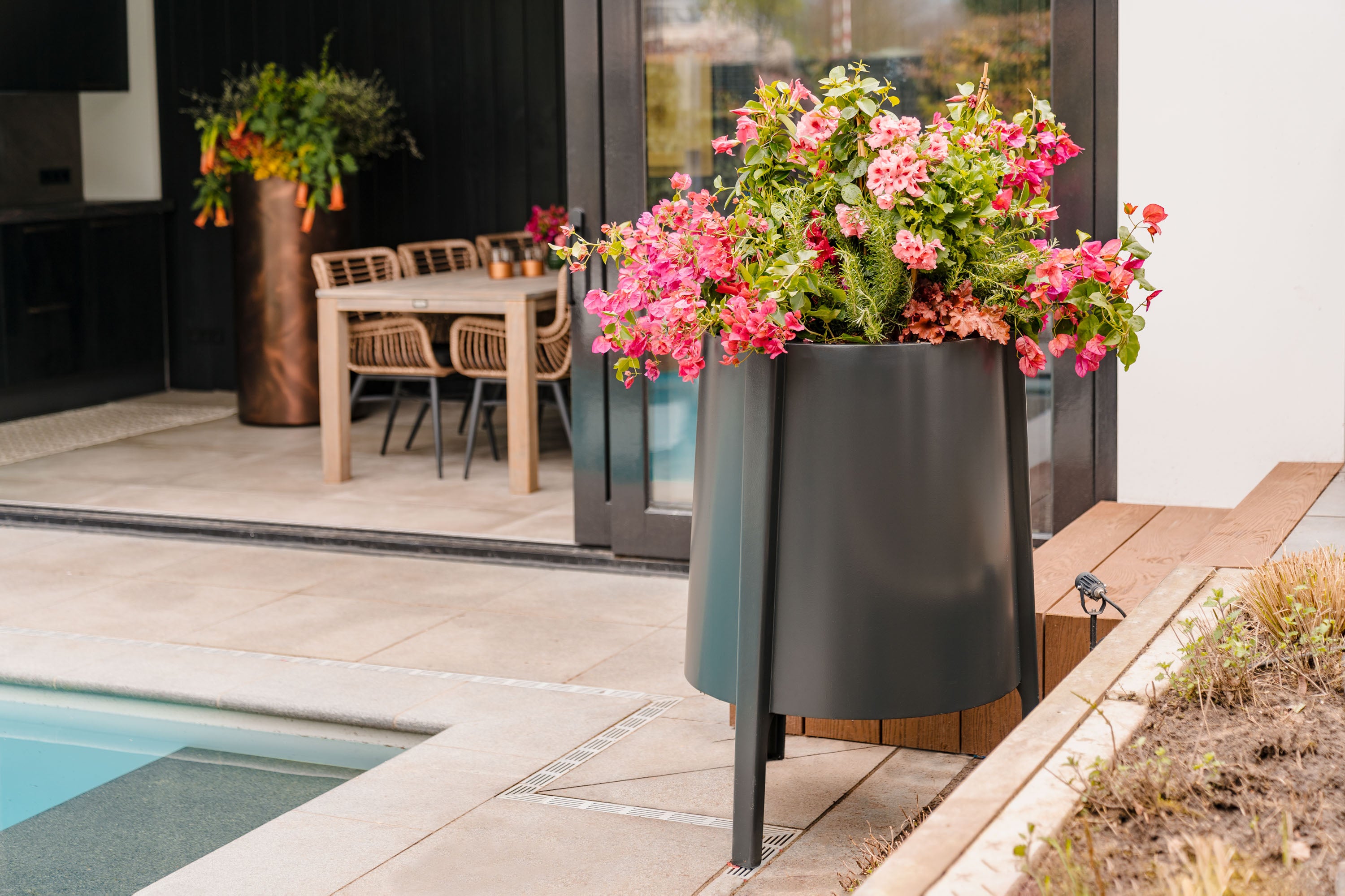 A large matte black cylindrical planter with tapered legs, filled with vibrant pink and red flowers and green foliage, set on a wooden deck next to a swimming pool.