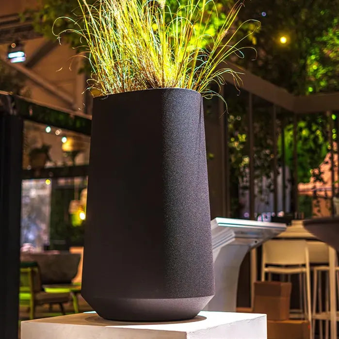 Rounded black tall garden planter. Planted with grasses and sited on a garden plinth.