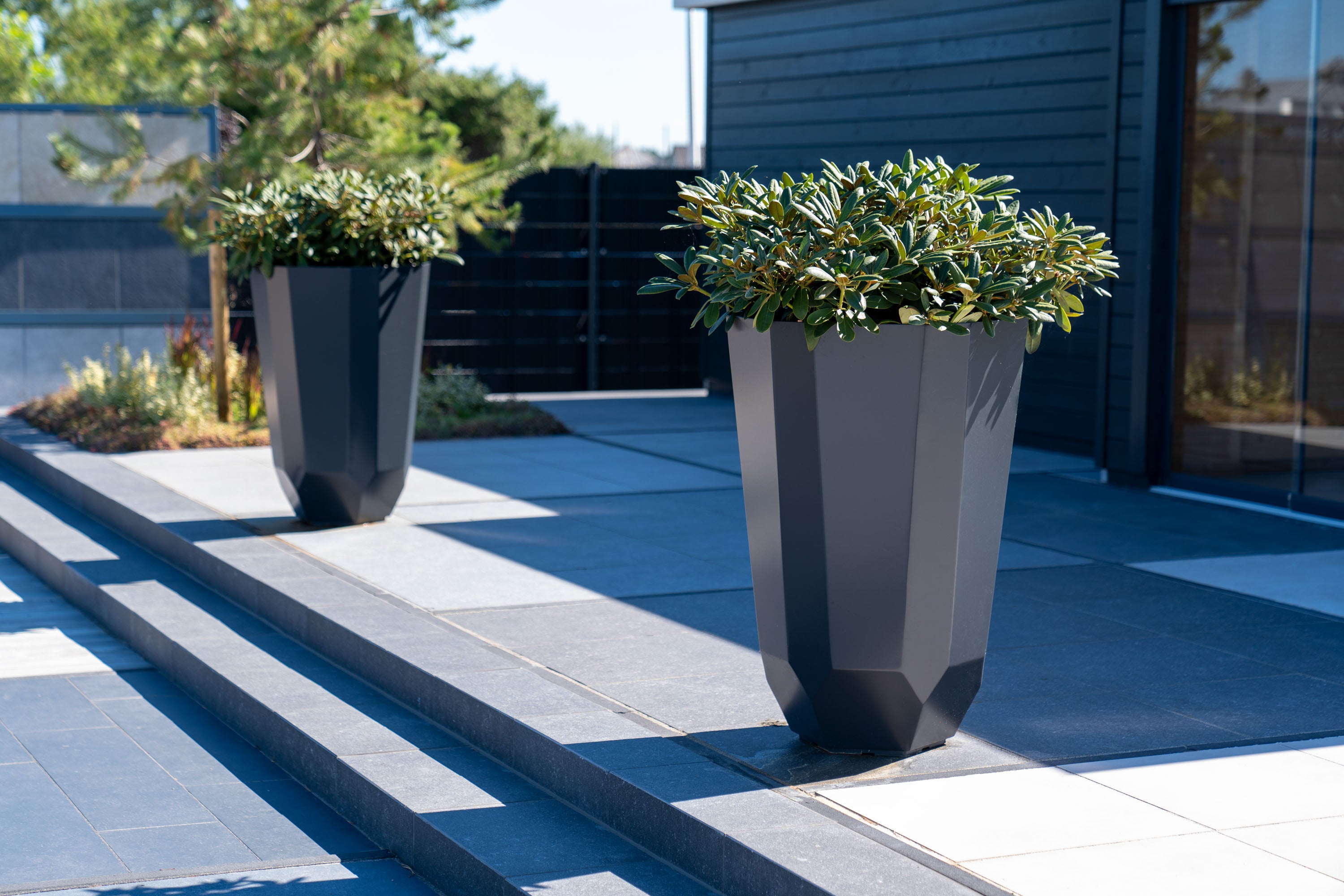 Two modern dark gray faceted planter boxes filled with green shrubs, placed on a paved patio in front of a contemporary house.