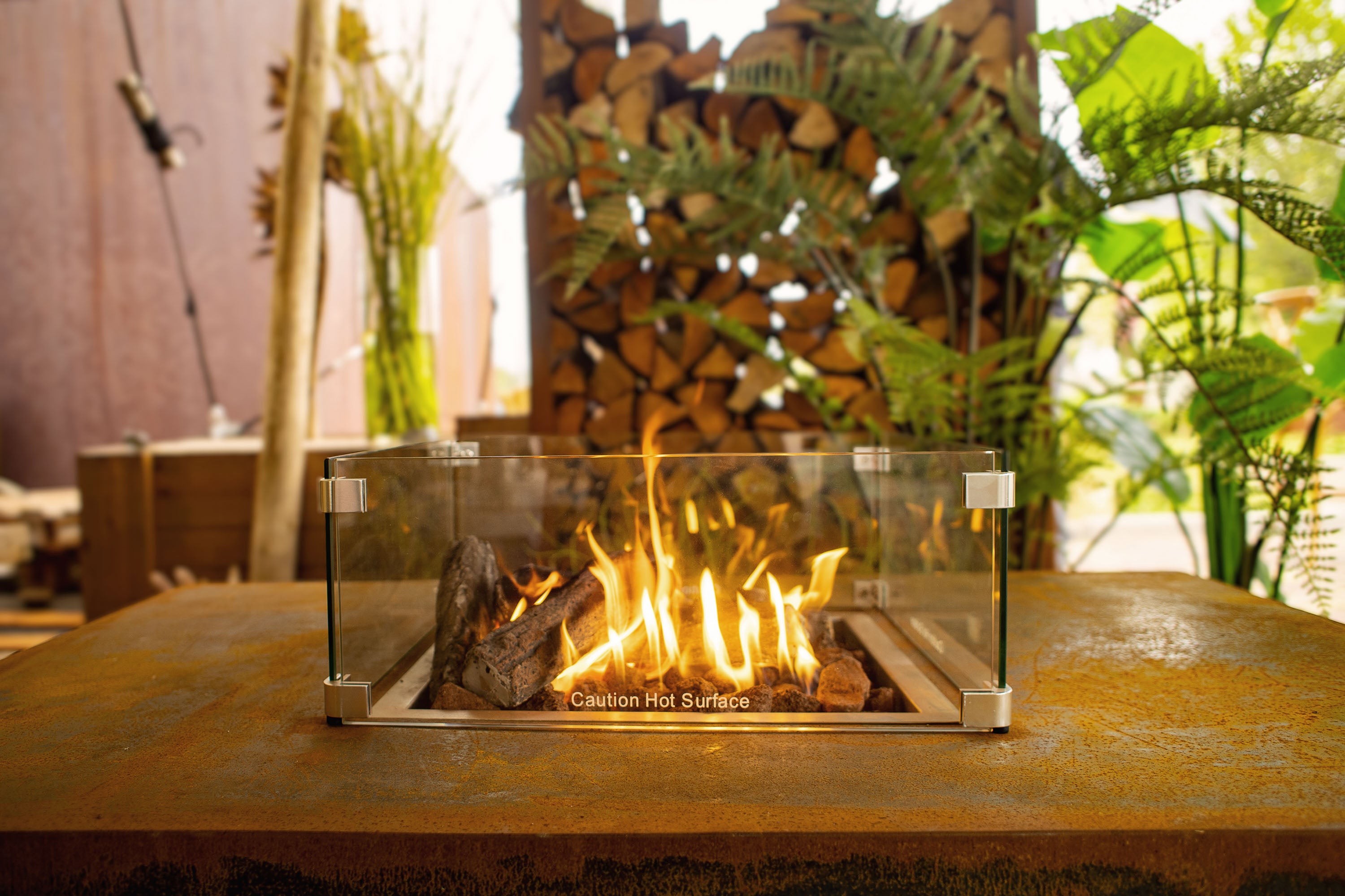 rustic finish fire table with clear central screen around lit fire in from of greenery