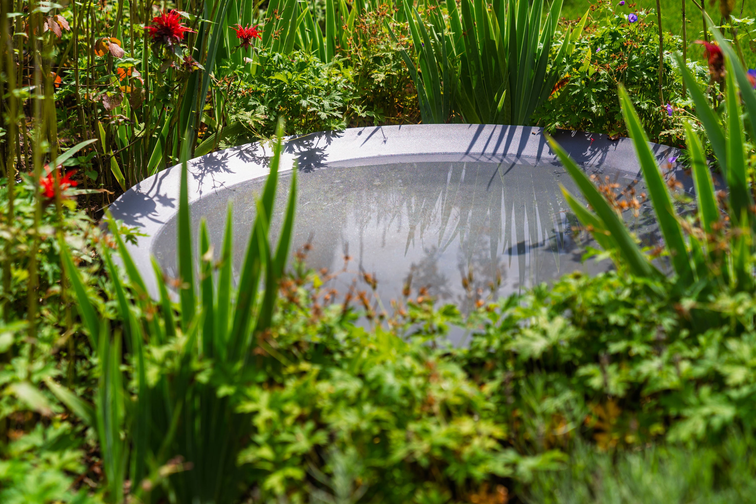 Round metal water feature in a garden with greenery and flowers