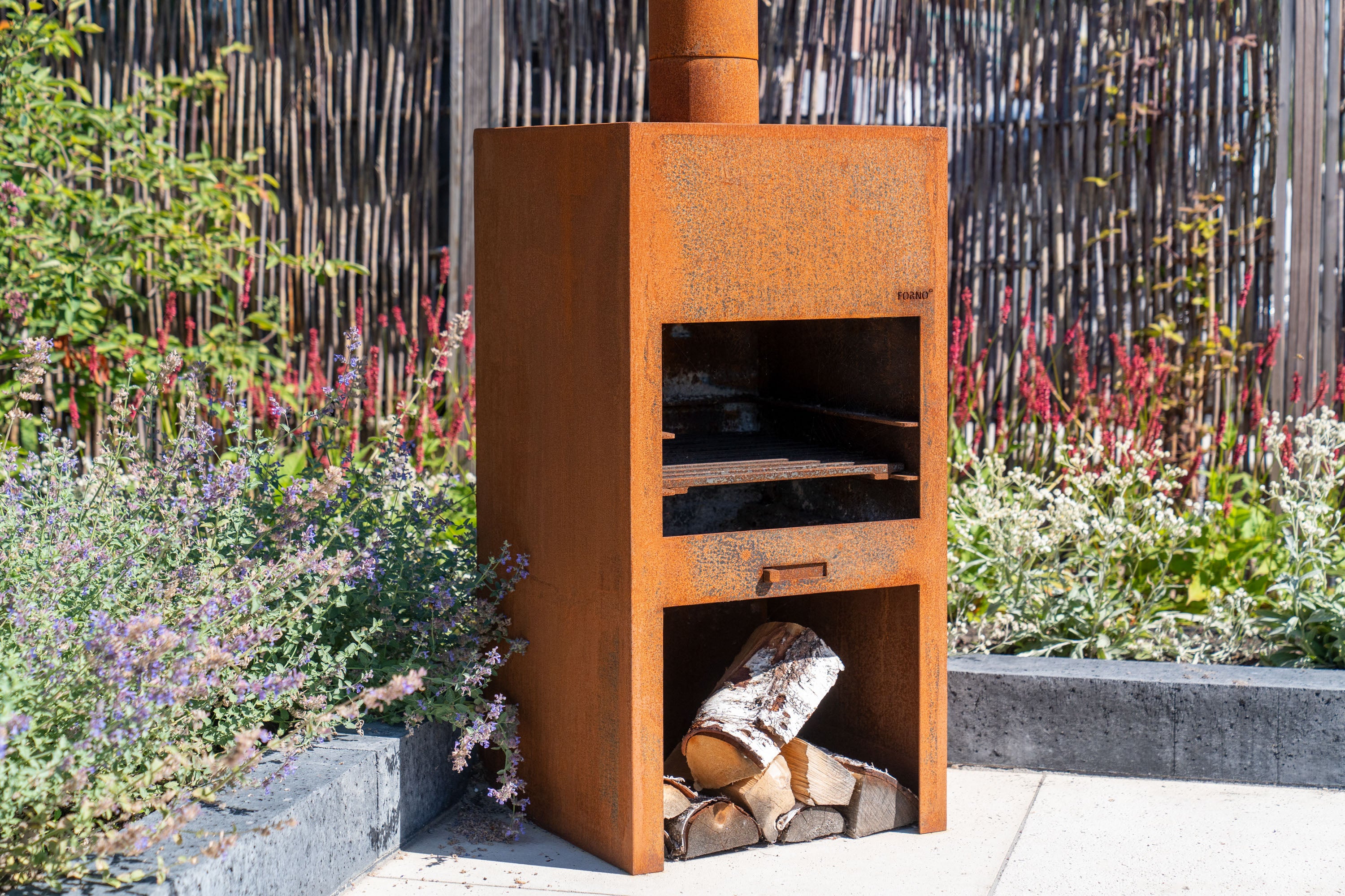 Rust-colored metal outdoor fireplace with logs inside, surrounded by plants and flowers.