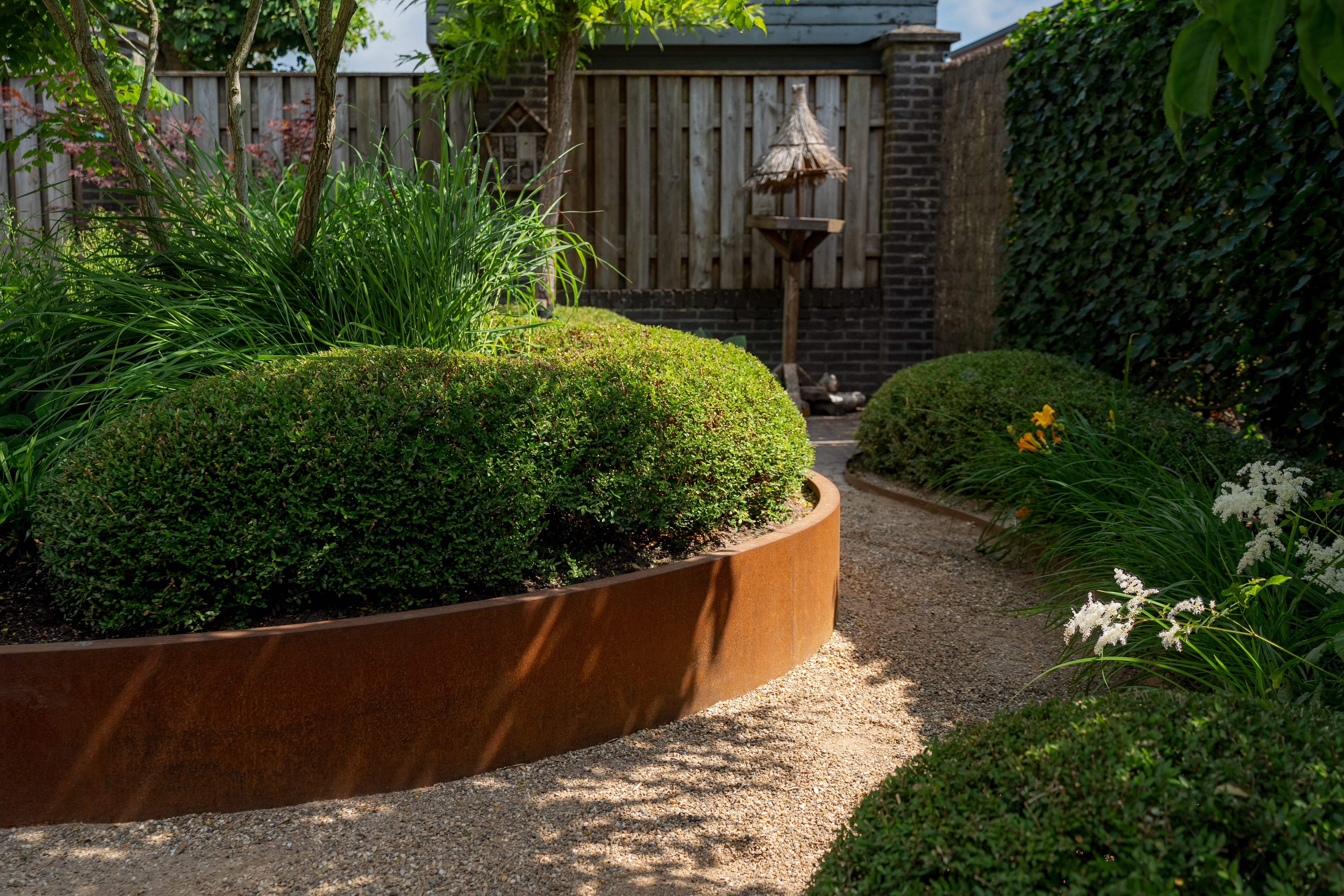 A landscaped garden pathway made of light gravel, bordered by rounded green shrubs in a rust-colored circular planter and dense greenery, with a wooden fence and brick wall in the background.