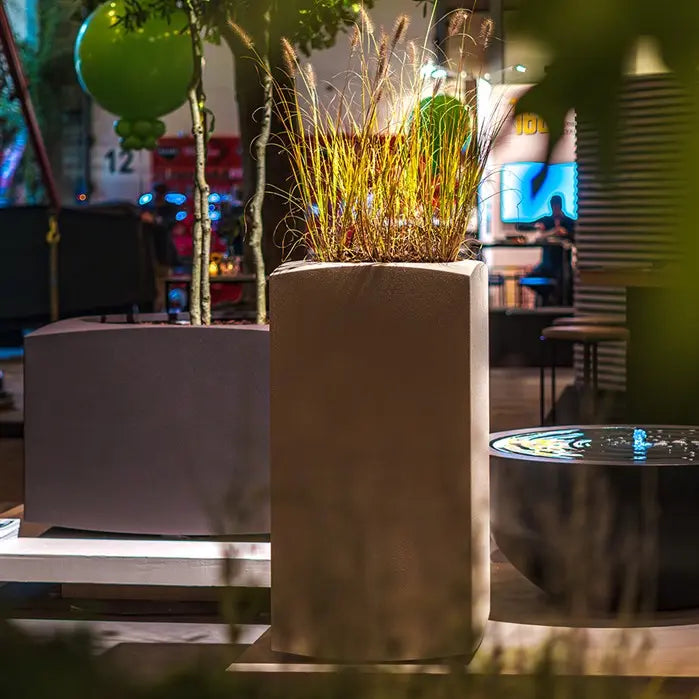 tall designer plant pot in cream. planted with grasses and situated indoors next to a water feature