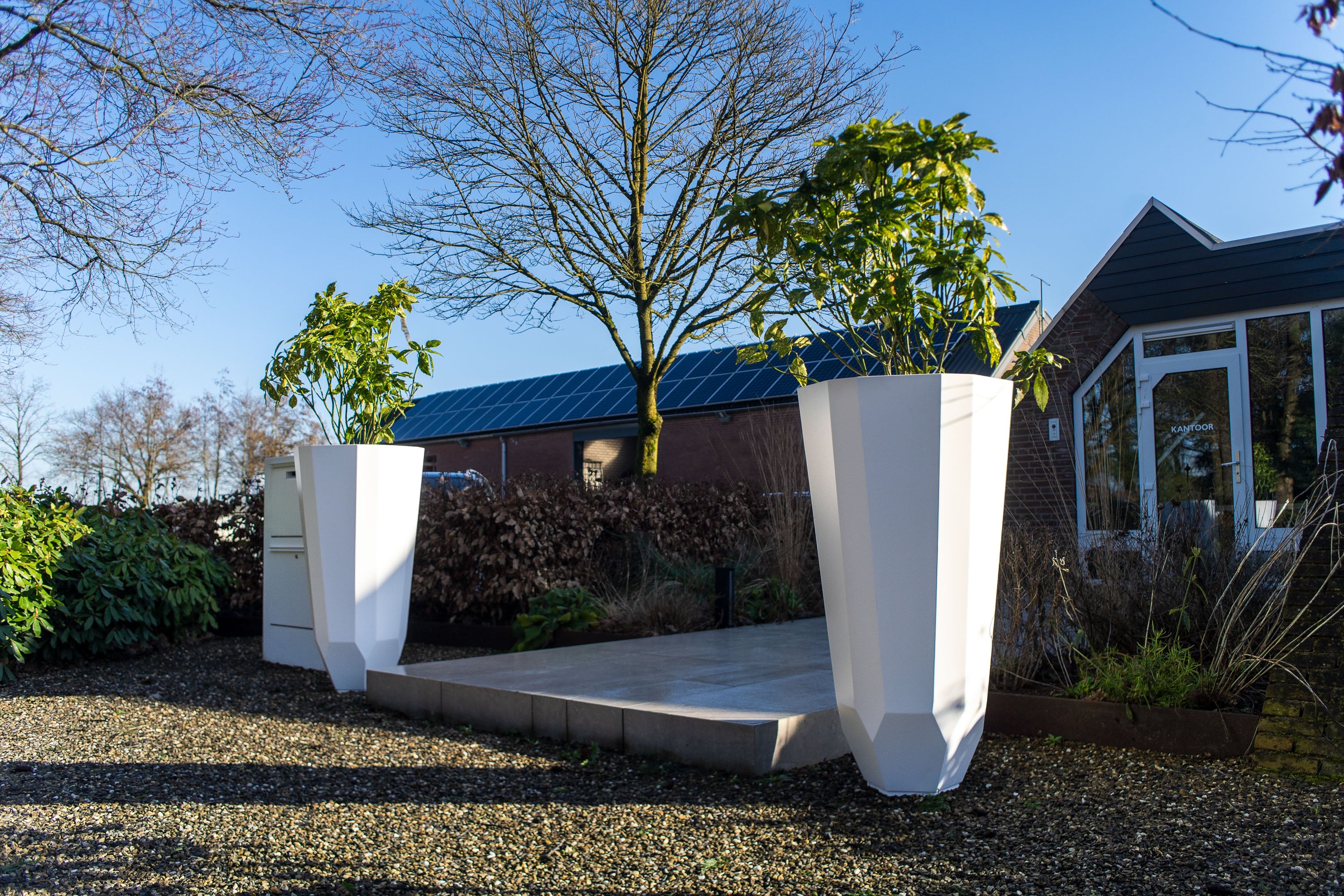 Two large white geometric planter boxes with green plants, placed on a concrete platform in an outdoor garden setting with gravel, shrubs, and a house in the background.