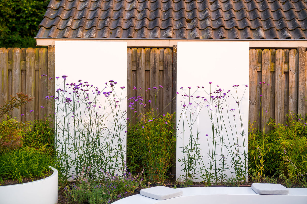 A white garden shed with a brown tiled roof, wooden doors, and tall purple flowers growing in front, set against a wooden fence with green plants and a white patio surface in the foreground.
