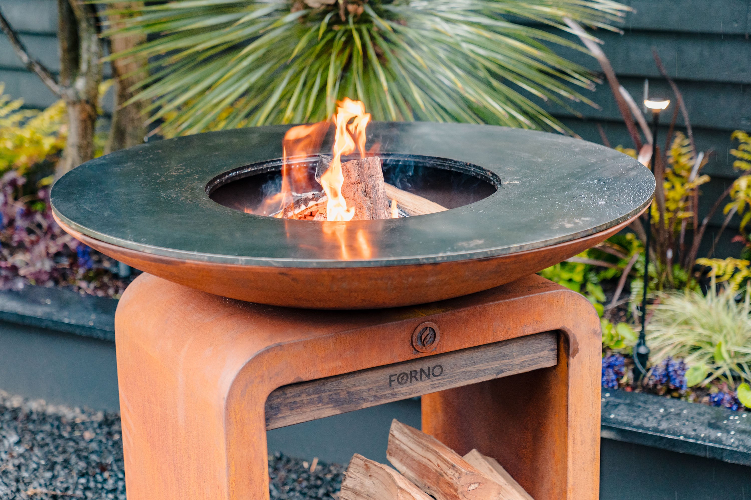 Outdoor fire pit with flames and 'Forno' branding in a garden setting