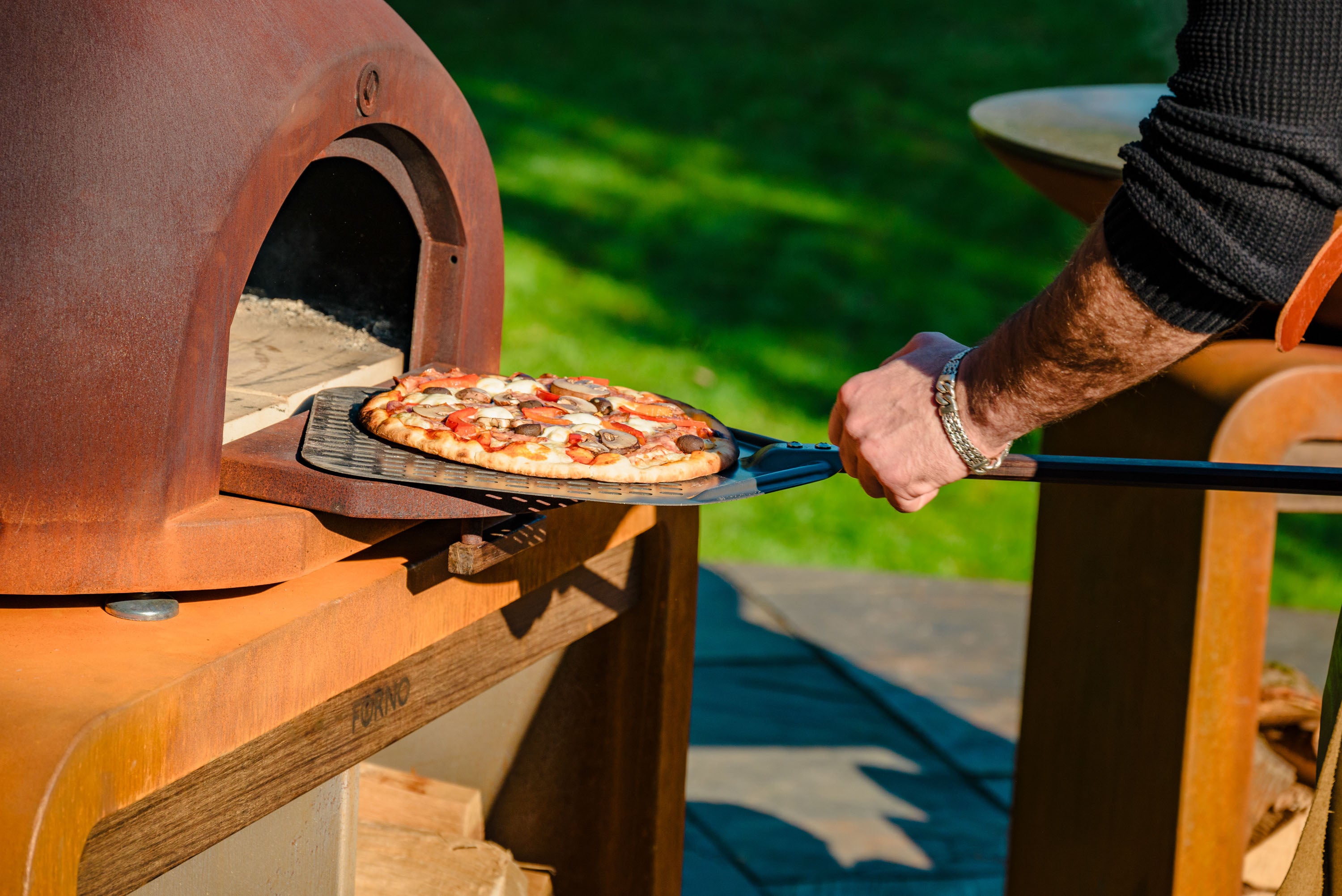 Person taking a pizza out of an outdoor brick oven with a scenic background.