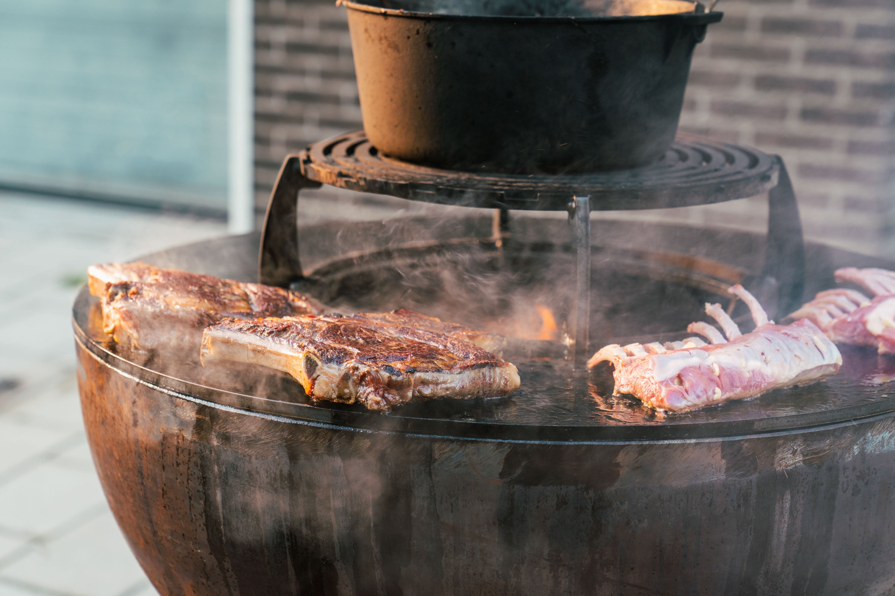 Steaks being cooked on a bbq grill with a pot above it.