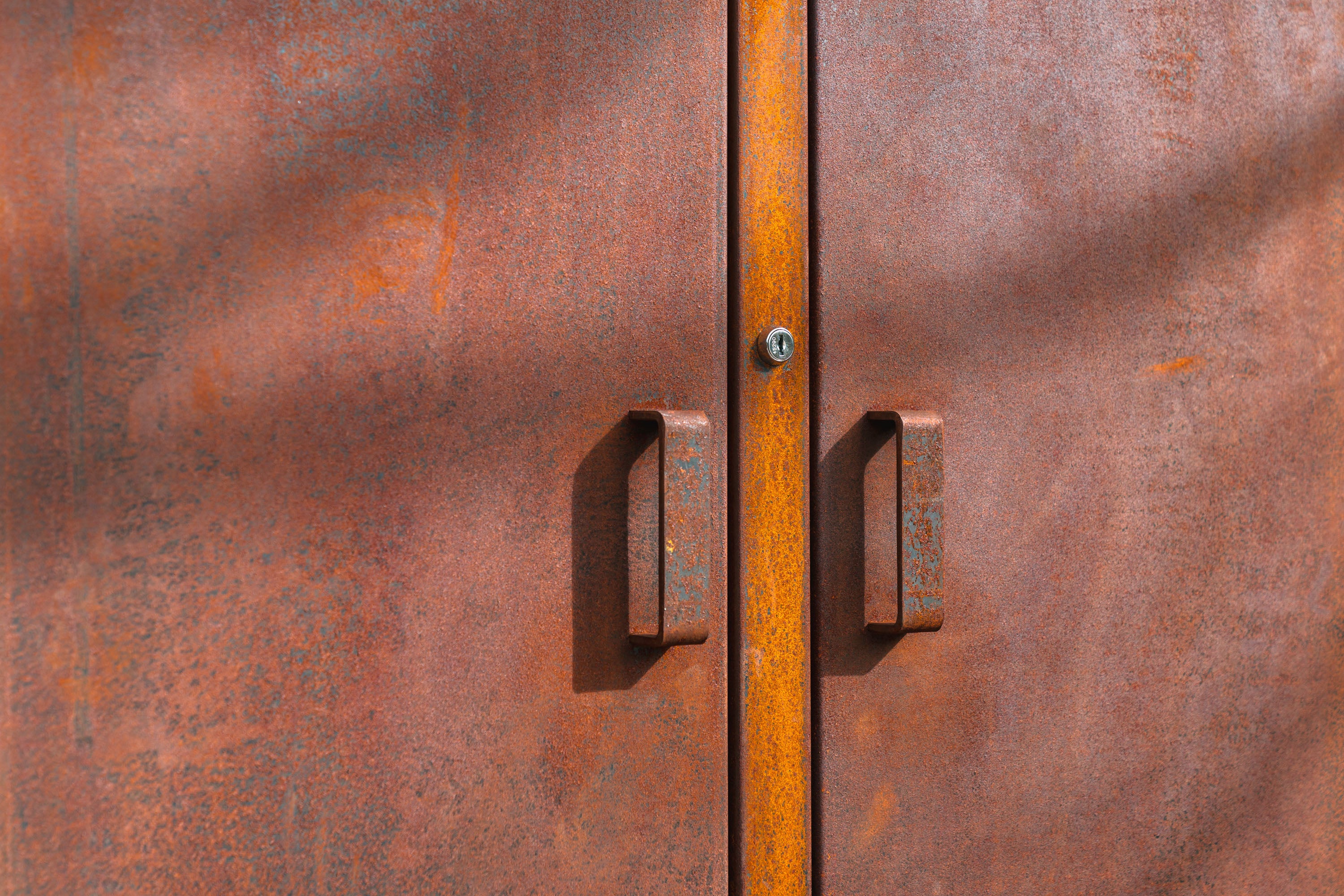 Close-up of a rusted metal double door with two vertical handles and a small round lock, showing a textured orange-brown surface with shadows.
