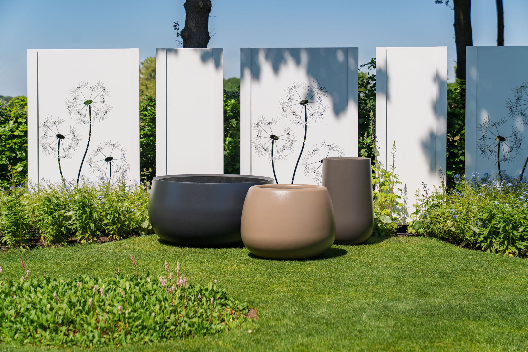 Three modern round outdoor planters in matte black, beige, and taupe, arranged on a green lawn in front of white panels with black dandelion illustrations.