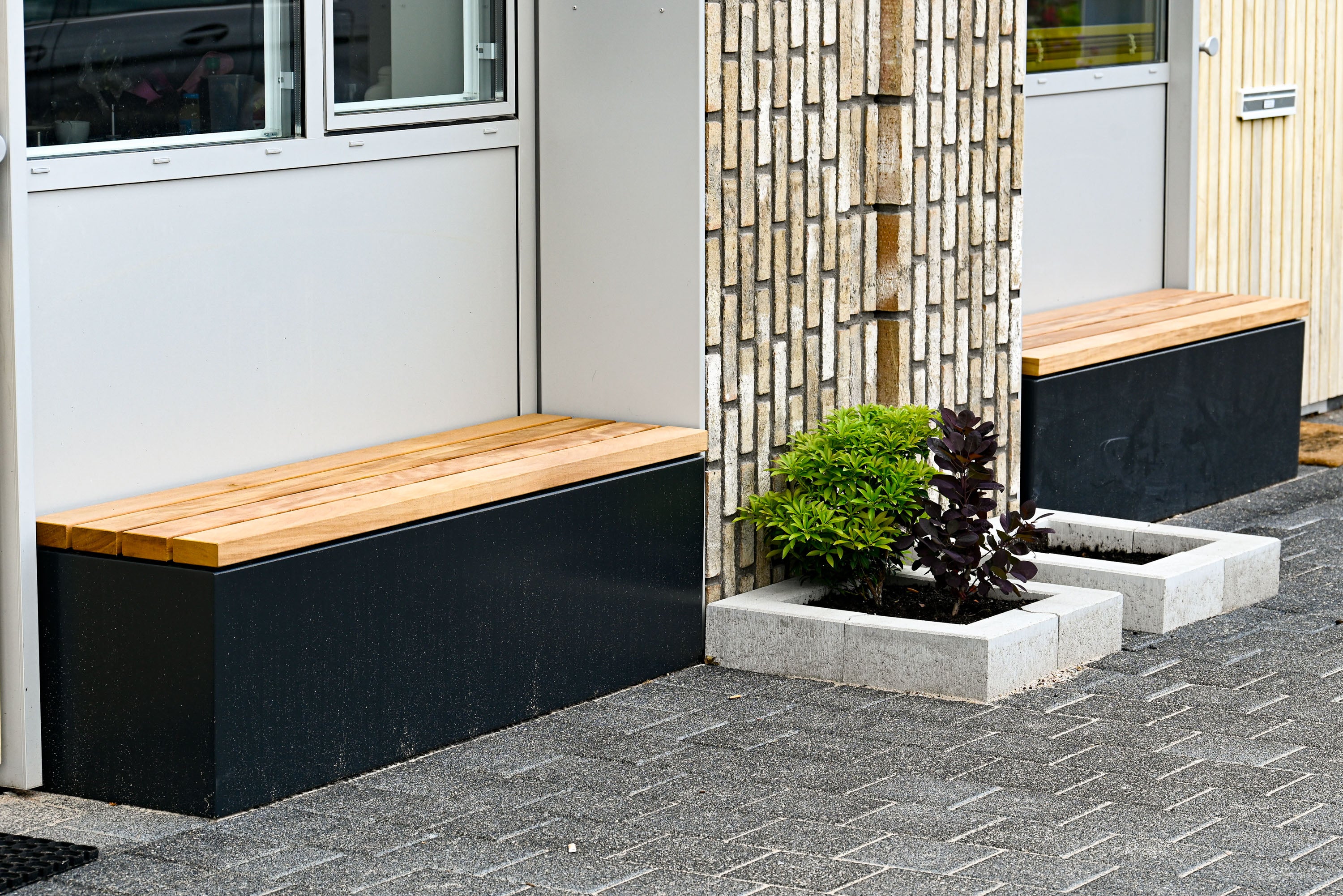 Modern outdoor wooden bench seating with black metal bases, installed beside a building entrance on a paved area with a planter in between.