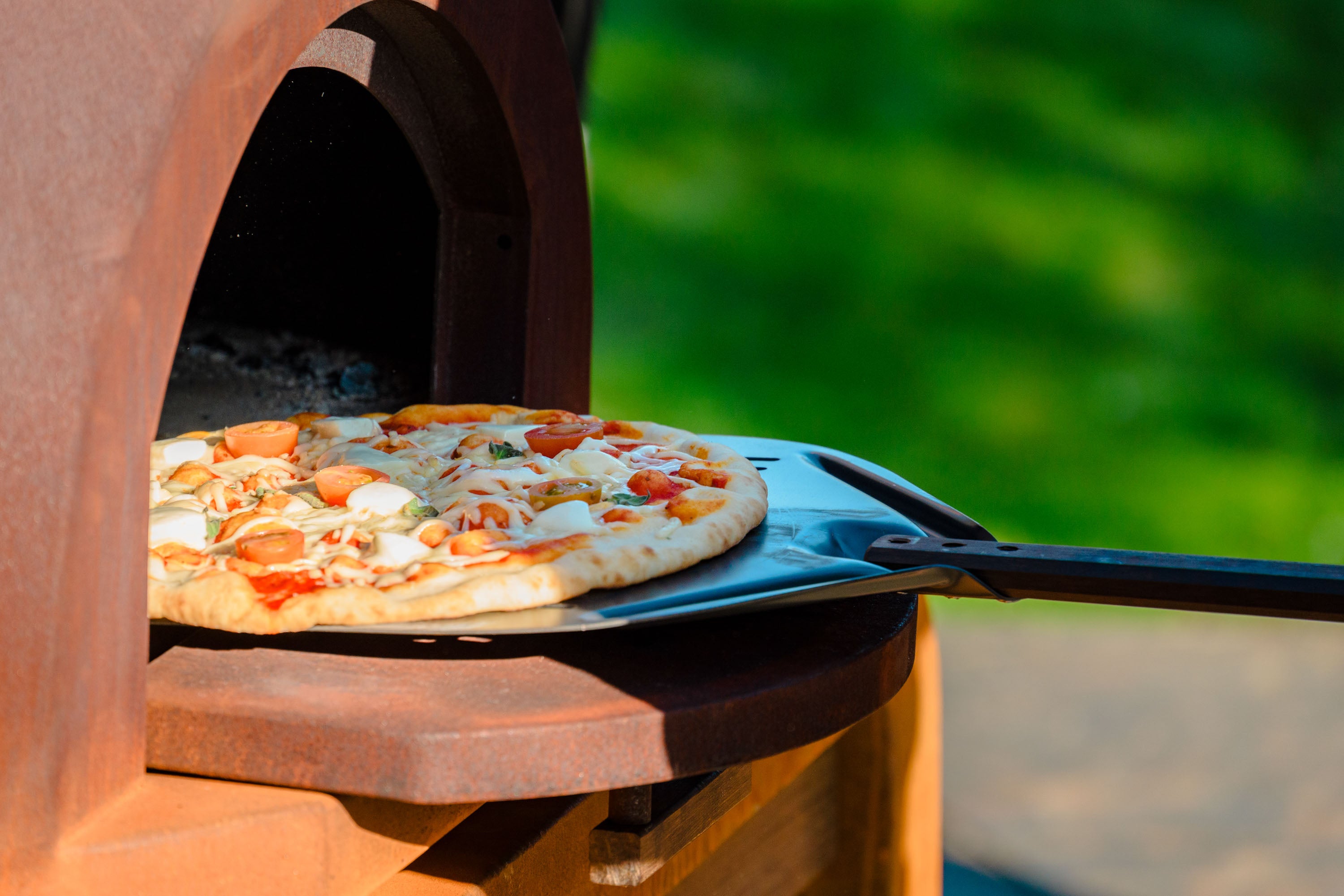Pizza being taken out of a wood-fired oven with a blurred green background
