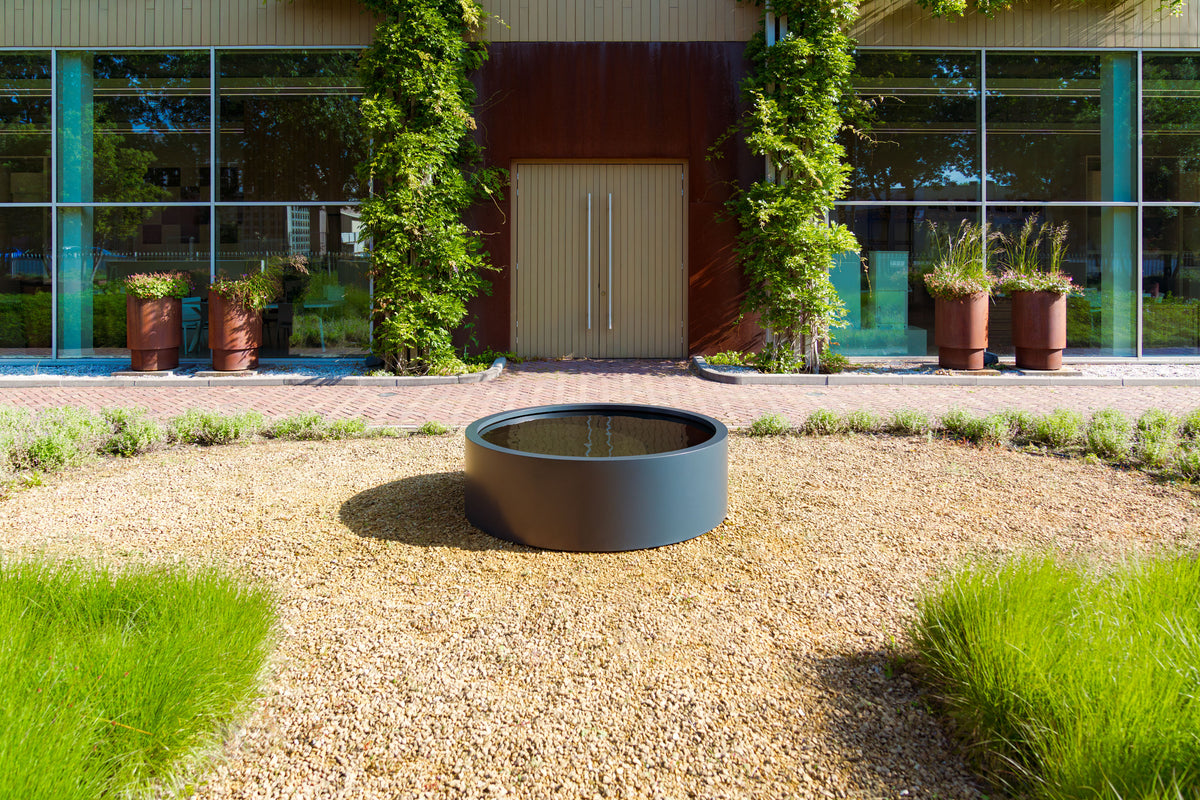 A round dark gray outdoor fire pit with water, set in a gravel landscape in front of a modern glass building with ivy-covered doors.