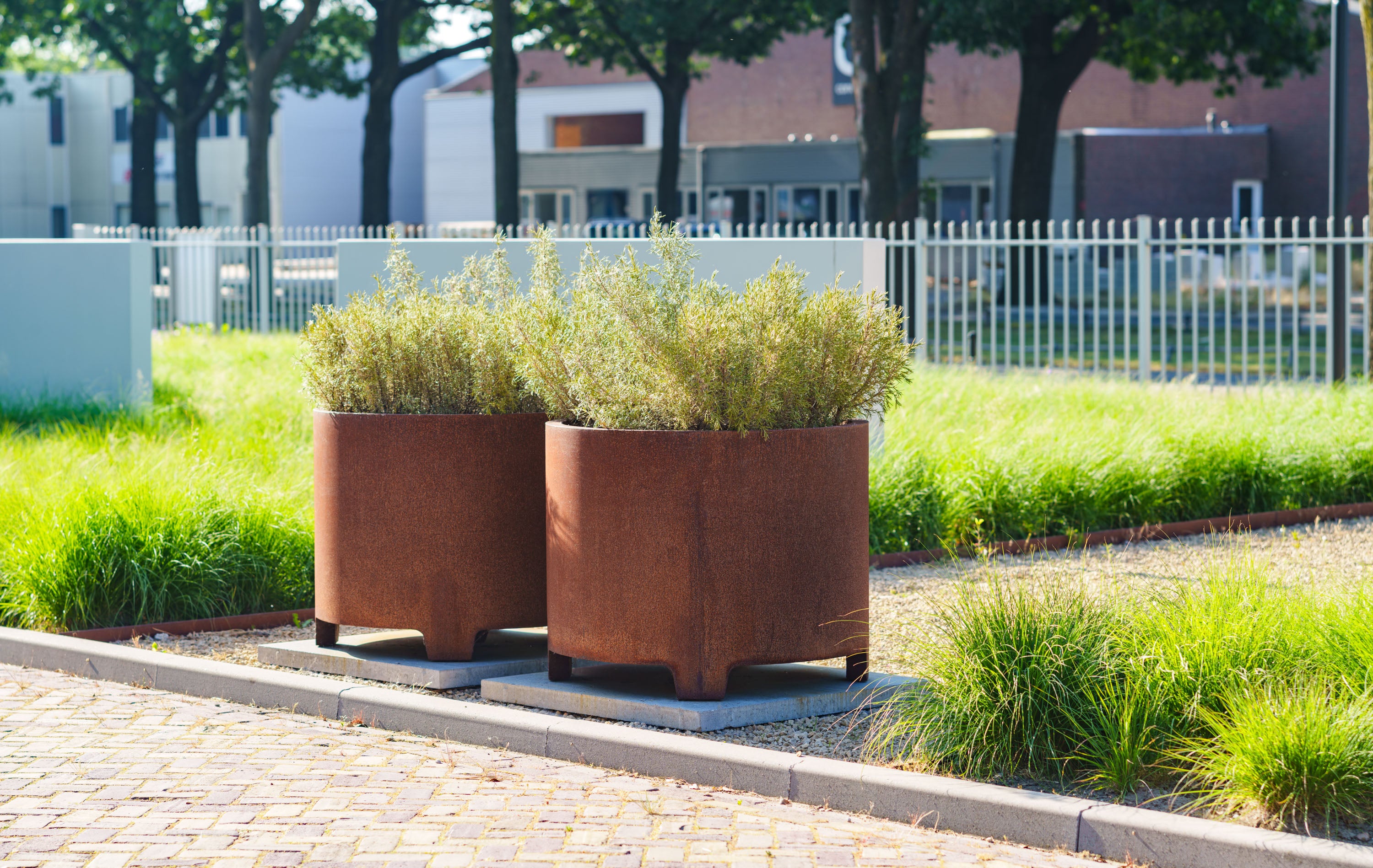 Two rust-colored planters with green plants on a sidewalk in an urban setting.