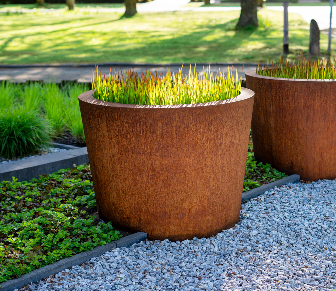 two round rusted planters, planted with grasses sitting between o gravel and plants