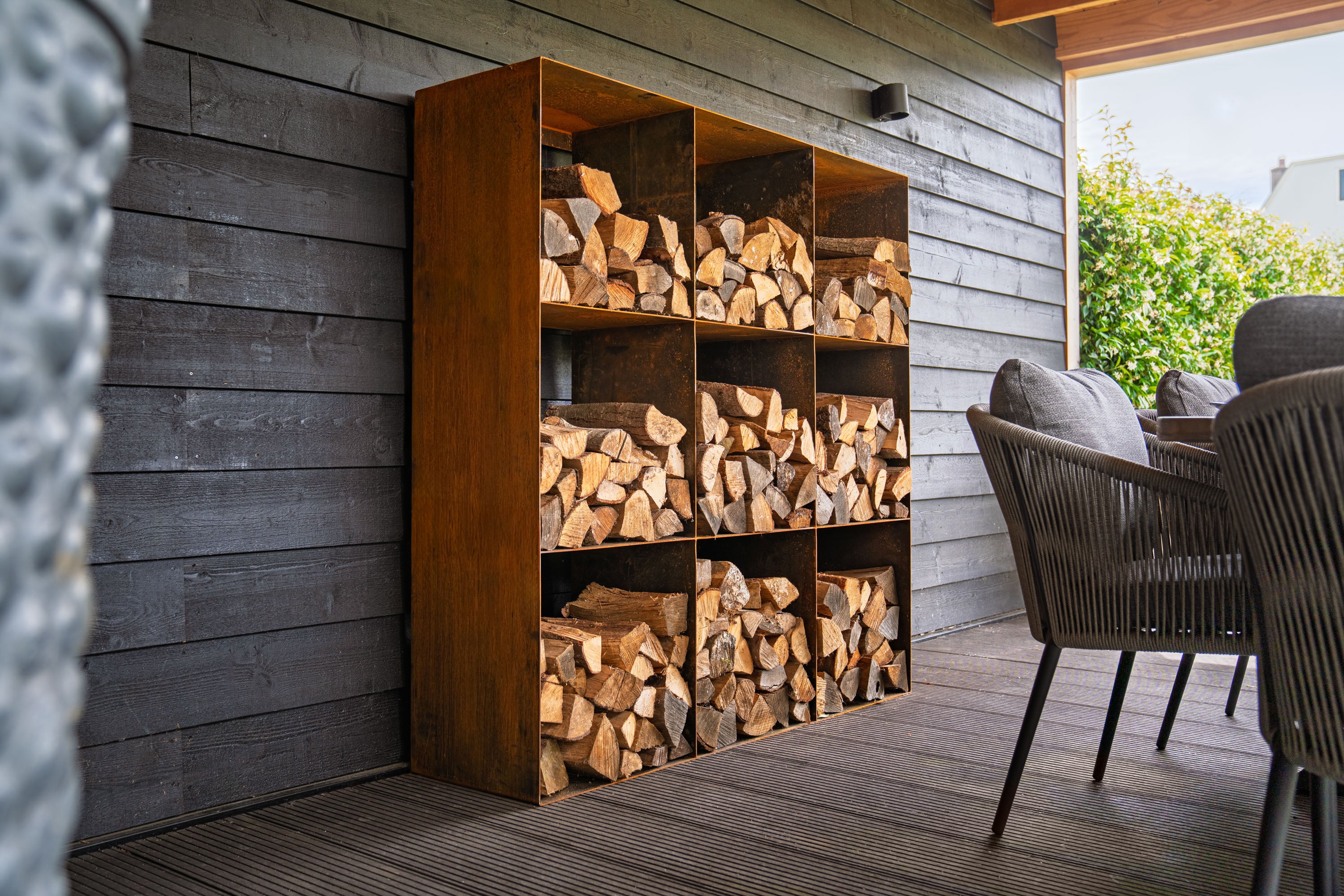 Wooden firewood storage unit against a wooden wall with outdoor furniture in the background.