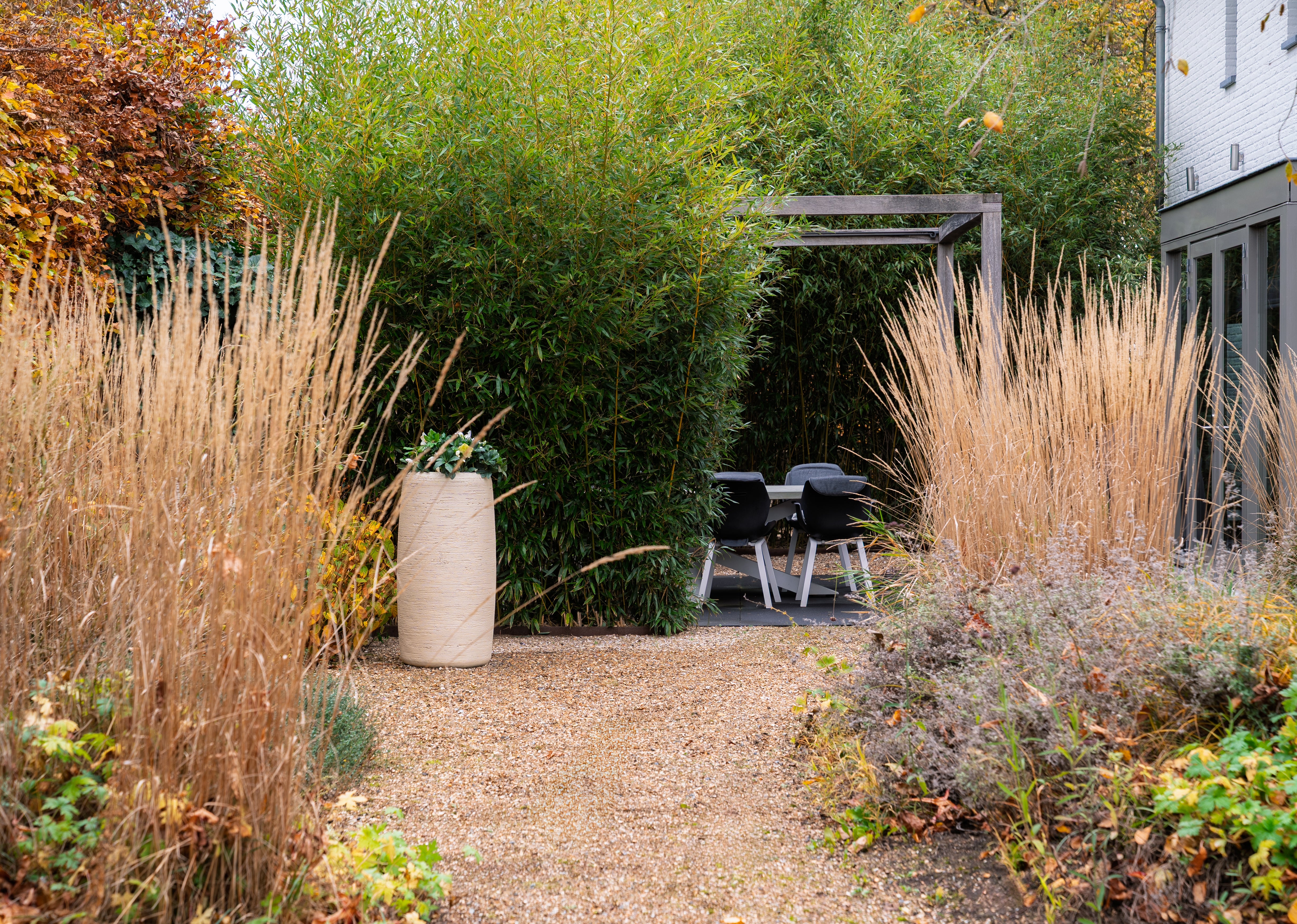 A landscaped garden pathway made of light gravel leads toward a small patio area with black outdoor dining chairs under a wooden pergola, framed by tall ornamental grasses, dense green bamboo, and shrubs. A large beige planter sits on the left side.