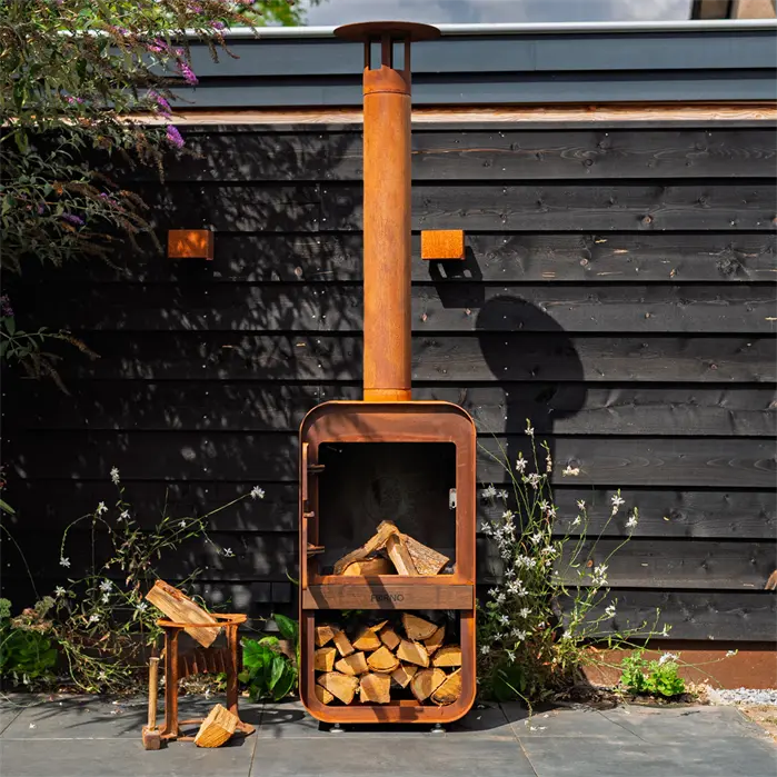 A rusted orange outdoor firewood stove with a tall chimney stack, filled with logs, set against a dark wood-paneled wall on a paved patio.