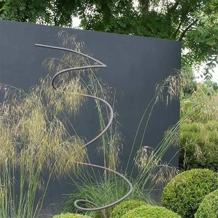 Metal spiral sculpture in a garden setting with a dark wall and greenery.
