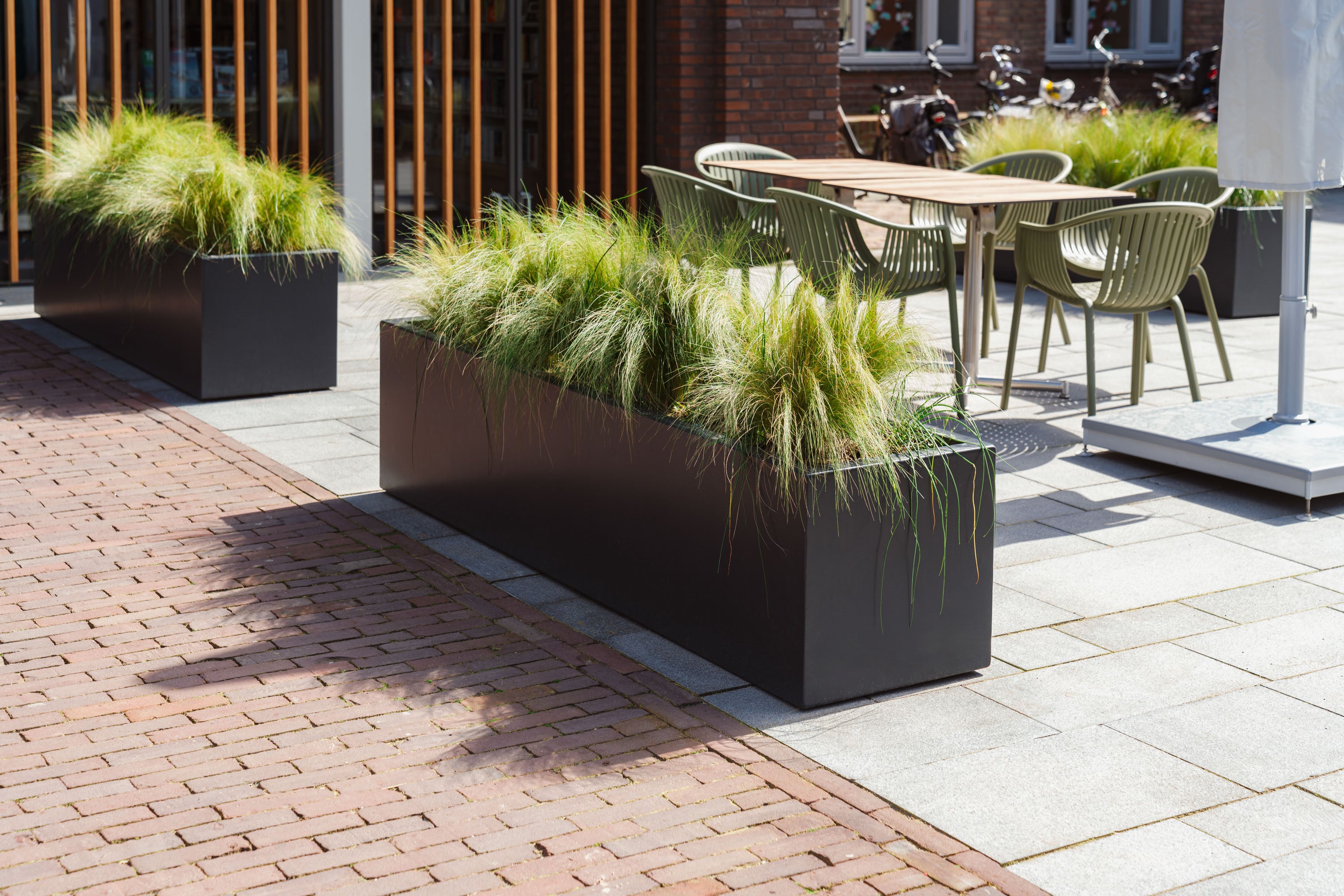 Outdoor seating area with green chairs and tables, surrounded by black planters with grass-like plants.