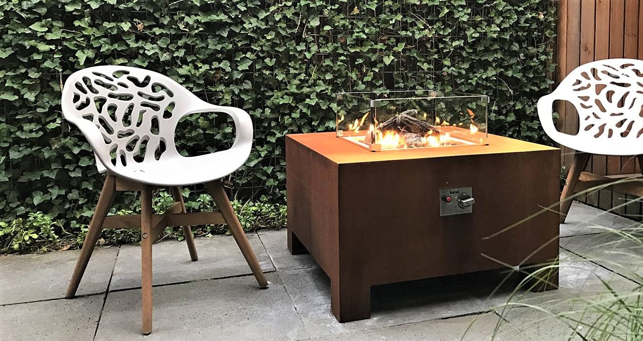 square rusted fire table with Perspex screen around lit central gas fire on paving in front of a green wall and next to two white chairs
