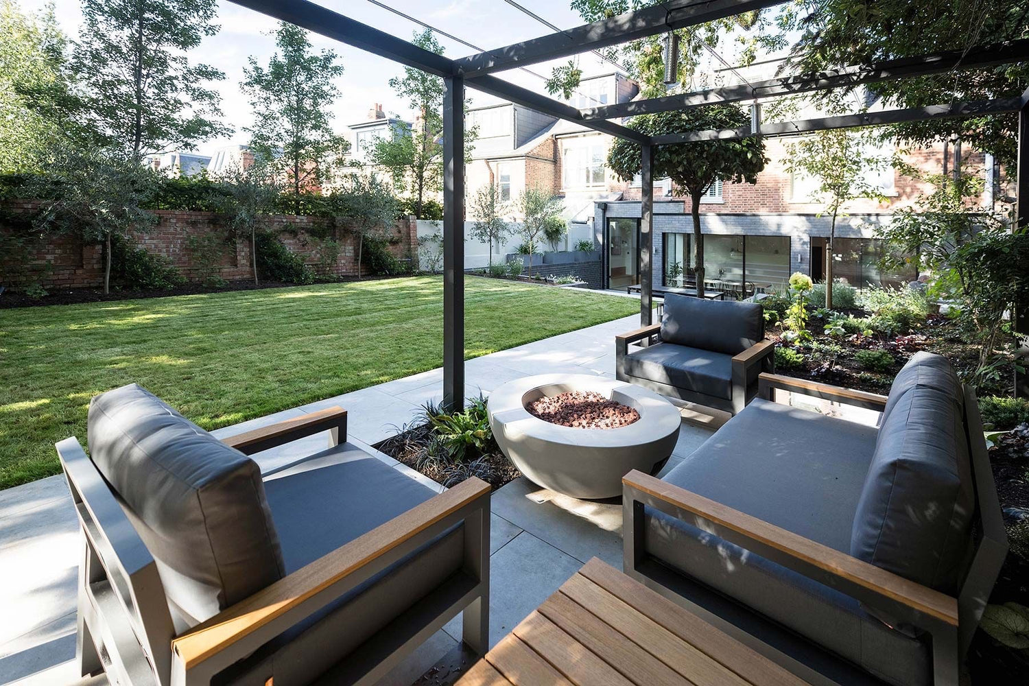 Black aluminium pergola over an outdoor seating area in a London town garden. There is a fire bowl and garden chairs and sofa.