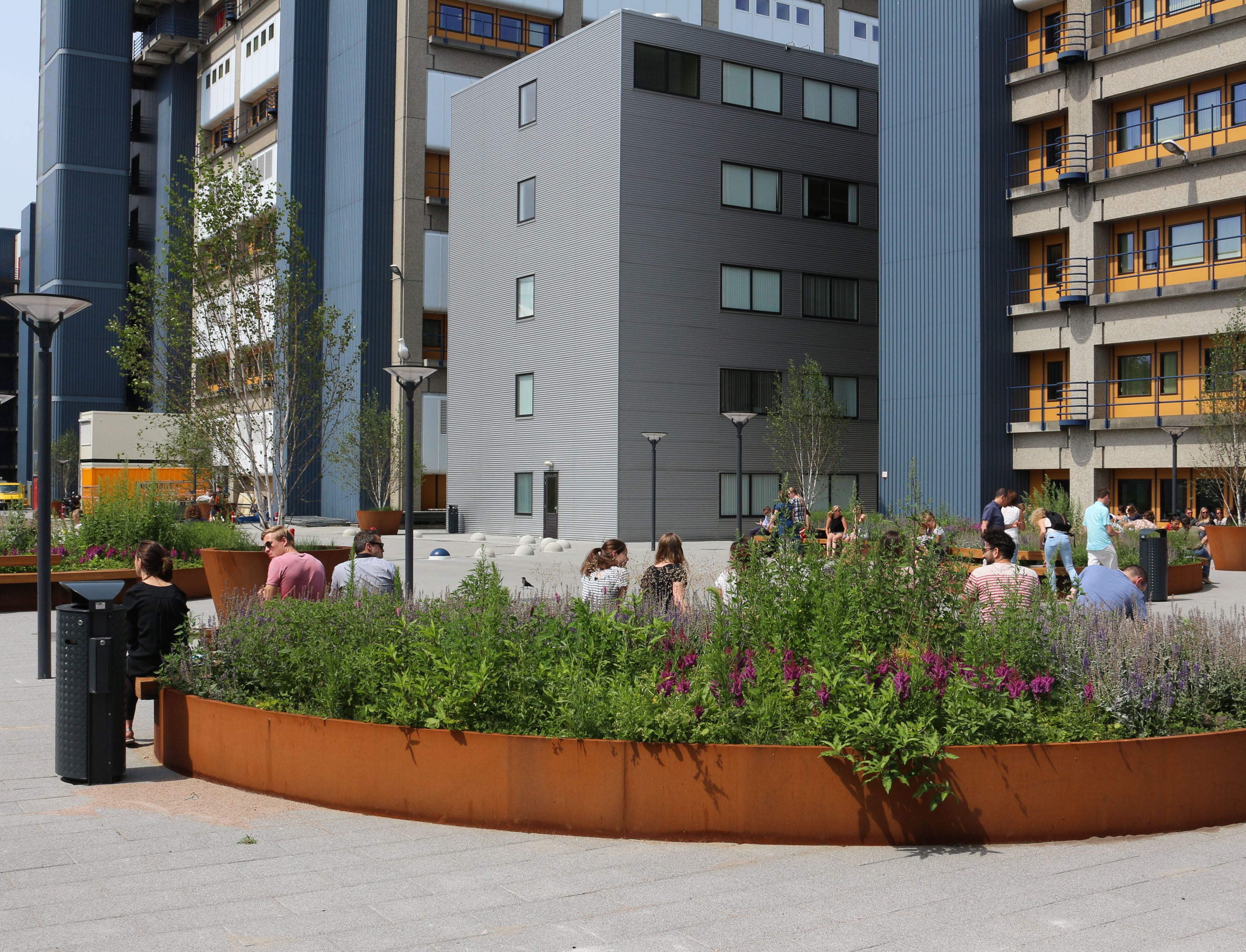 Outdoor urban plaza with modern gray and brown buildings, a curved rust-colored planter bed filled with green plants and purple flowers, and several people sitting and walking in the background.