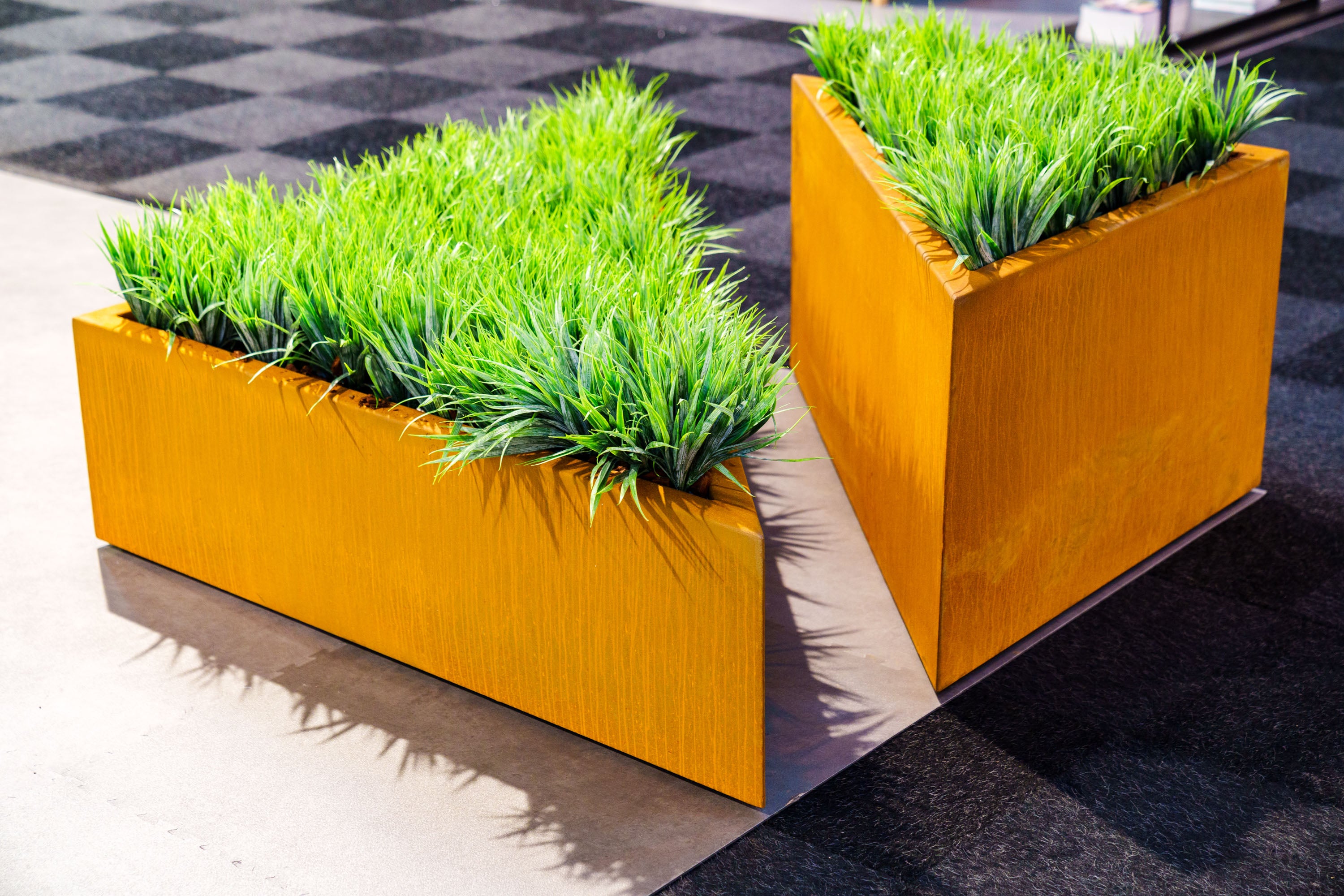 Two modern orange corten planters with green grass-like plants on a checkered pavement background.
