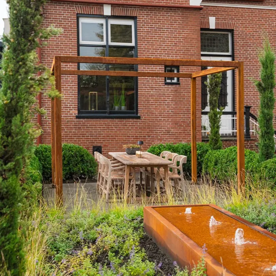 Outdoor patio area with wooden pergola, table, chairs, and water feature in front of a brick building.