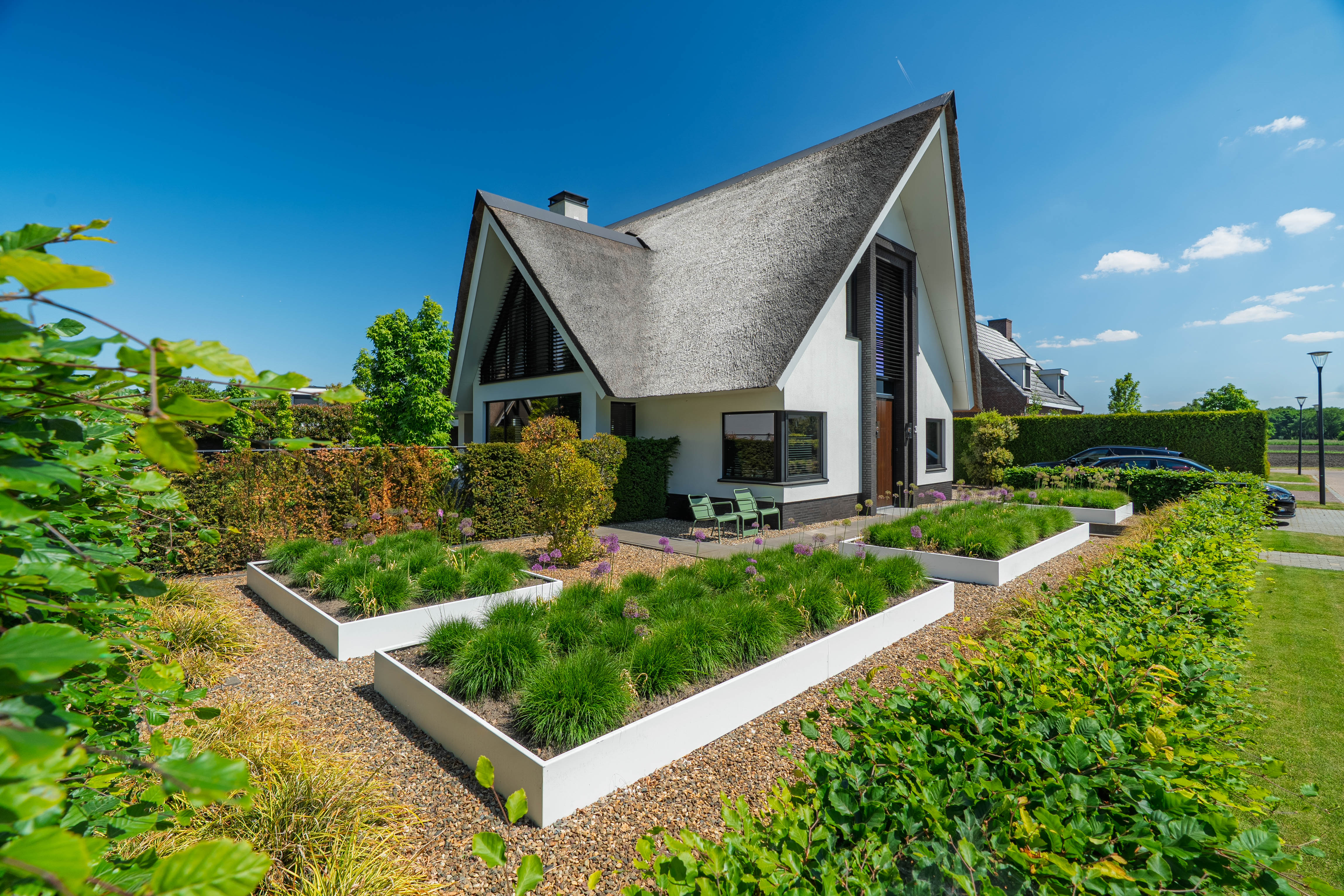 A modern single-story house with a steep thatched roof, white walls, and large windows, set in a landscaped garden with raised planters, gravel paths, and a blue sky background.