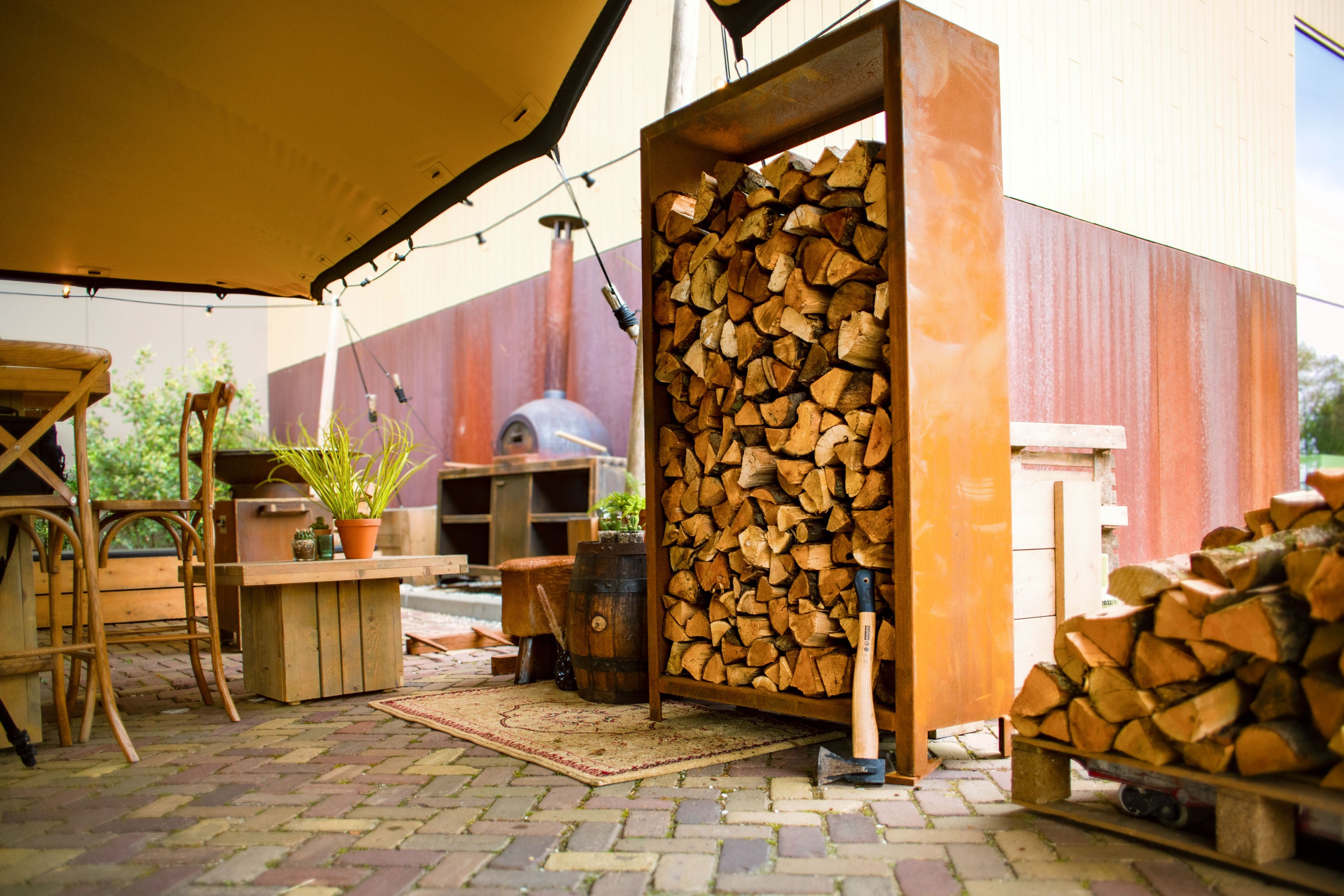 Wooden firewood storage unit on a patio with outdoor furniture and a building in the background.