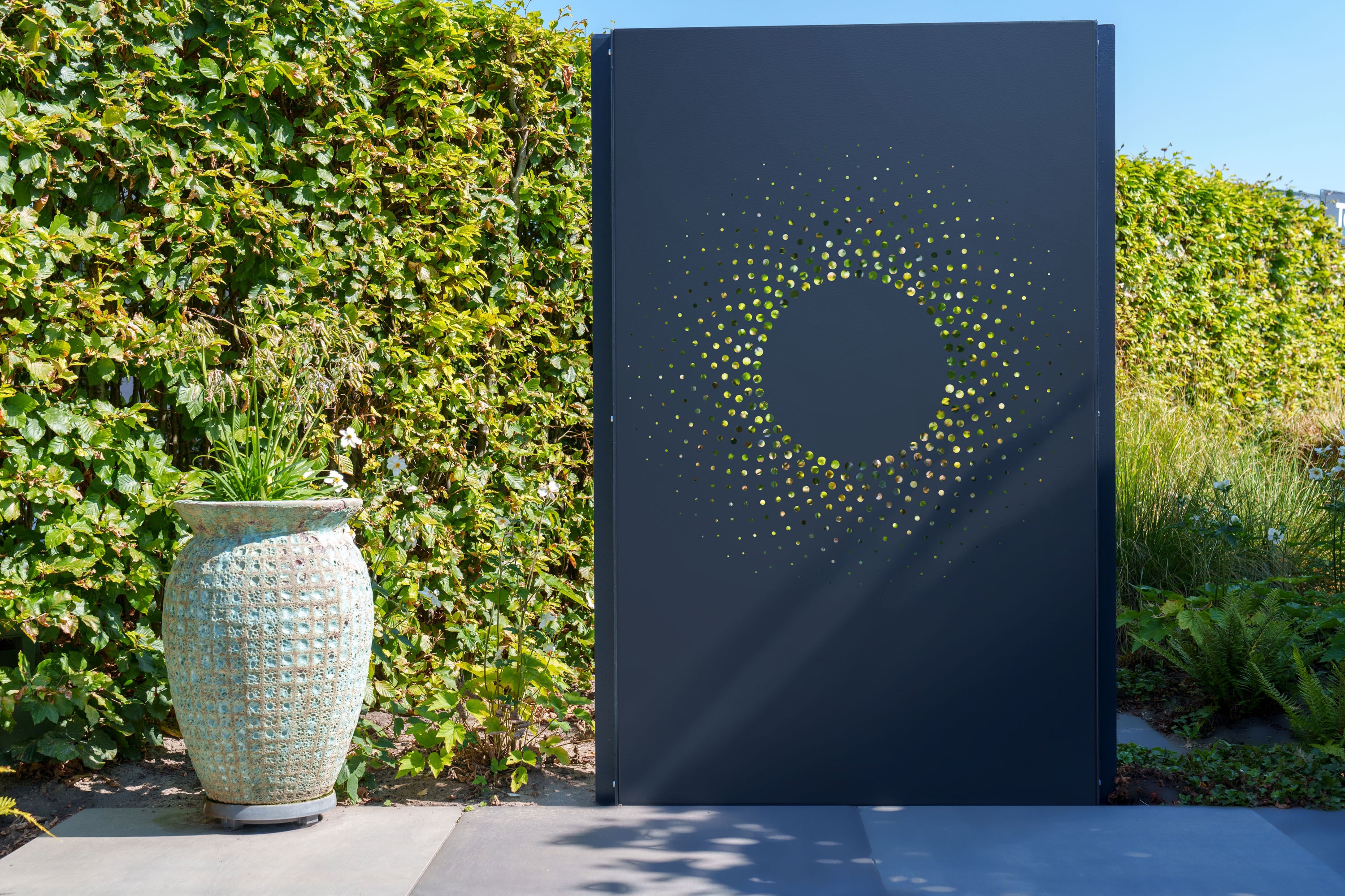 A tall, dark charcoal metal garden screen panel with a circular starburst cutout pattern, standing outdoors next to a large textured ceramic planter against a green hedge background.