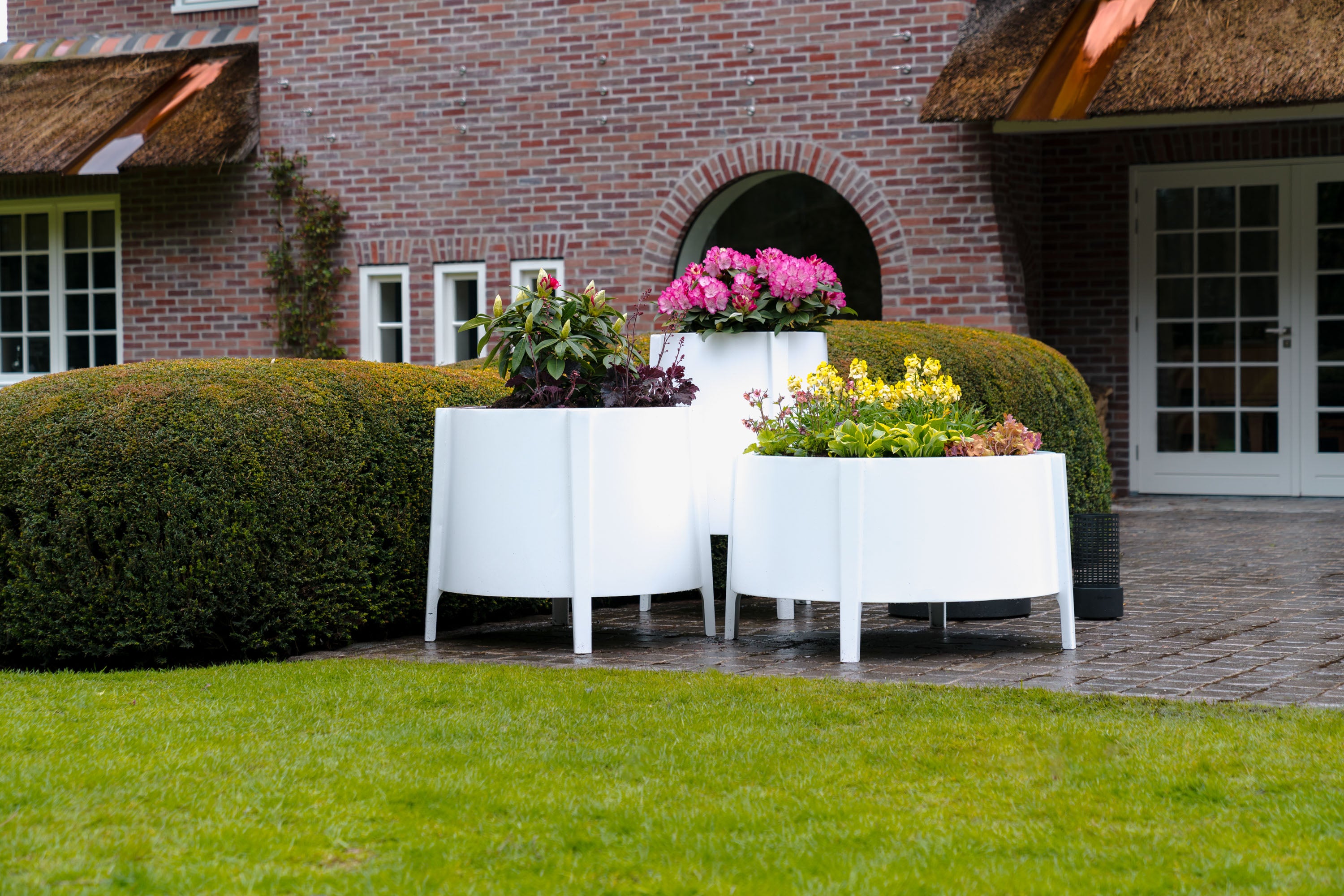 Three modern white curved planter boxes filled with colorful flowers, set on a stone patio in front of a brick building with manicured hedges and a green lawn.