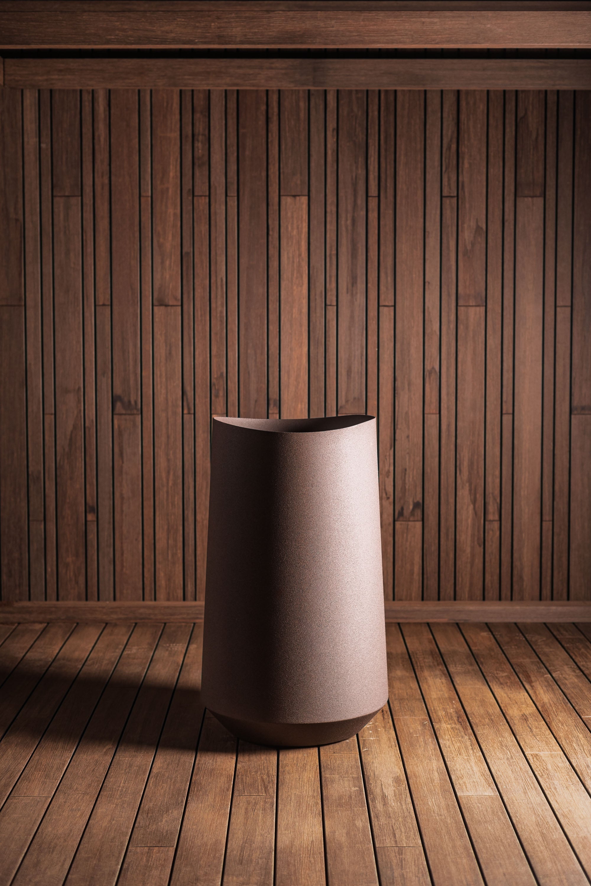 A modern cylindrical floor lamp with a textured taupe fabric shade stands on a wooden plank floor against a matching dark wood slat wall.