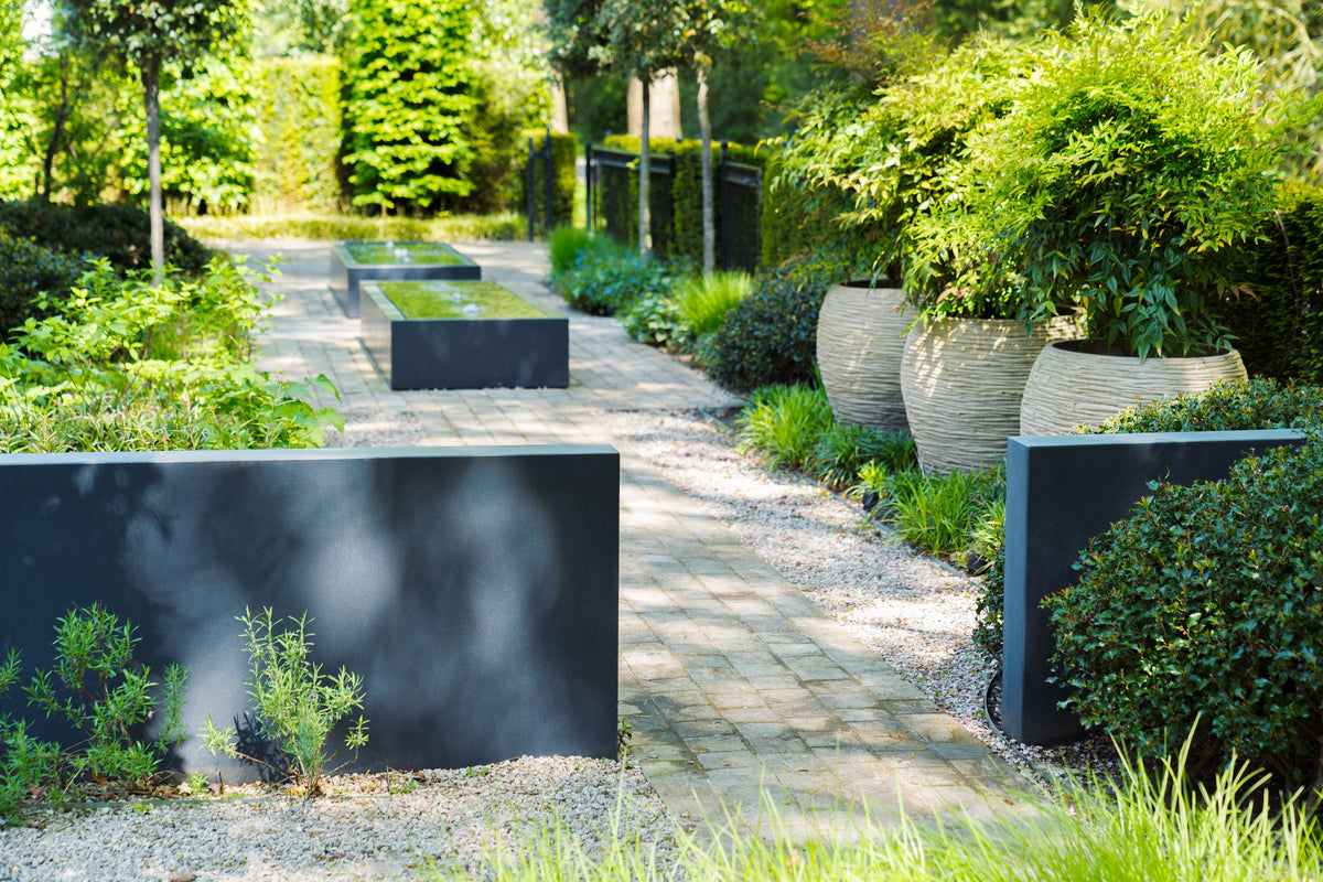 Modern minimalist garden pathway with black rectangular planter boxes, large beige textured planters filled with green foliage, and gravel paths surrounded by lush landscaping.