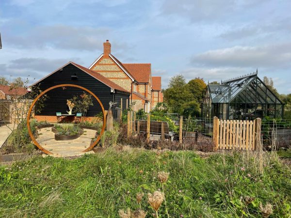 Garden with a greenhouse, wooden walkway, and circular metal sculpture.