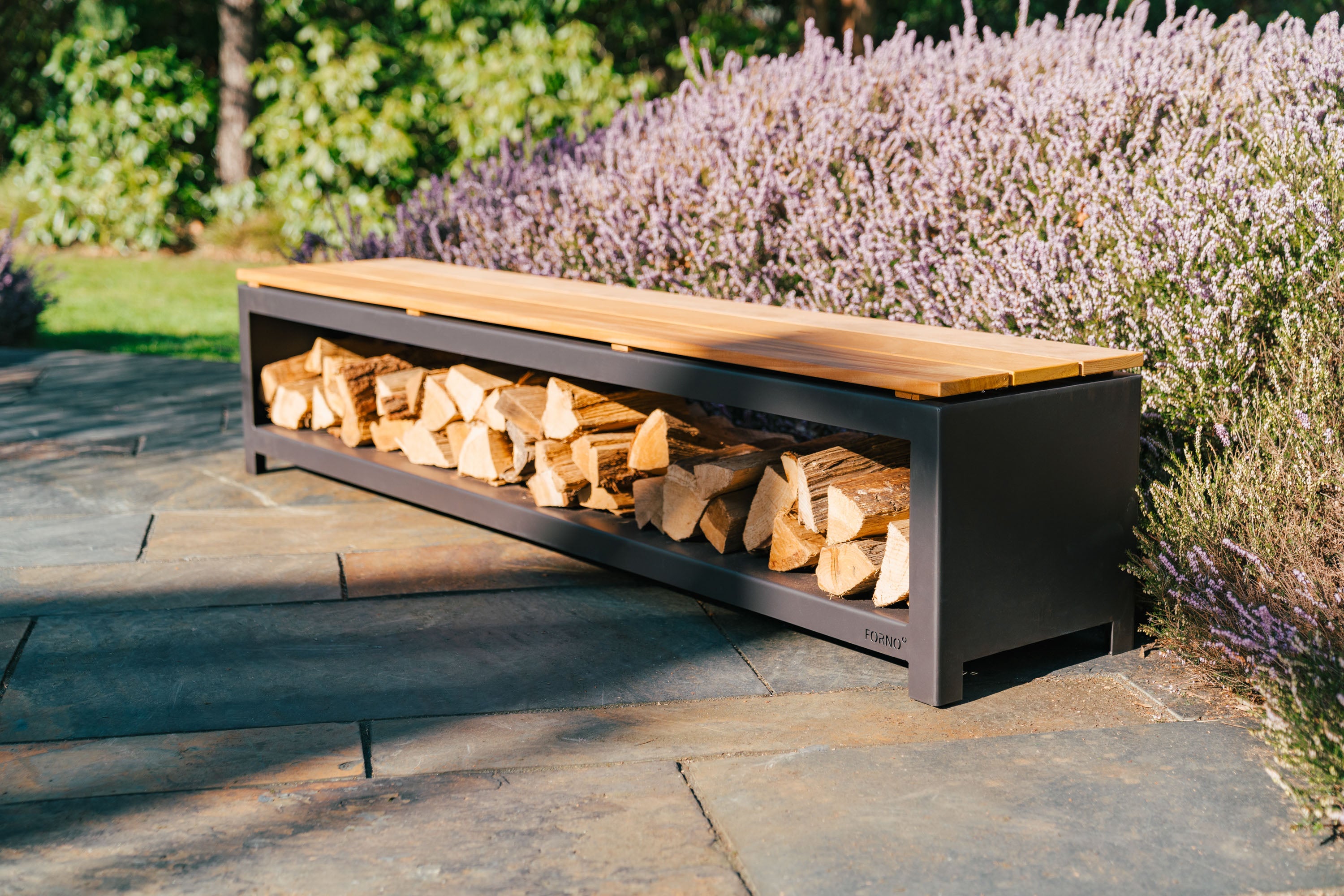 black wood storage bench filled with wood with wooden top on paving next to flowering bush