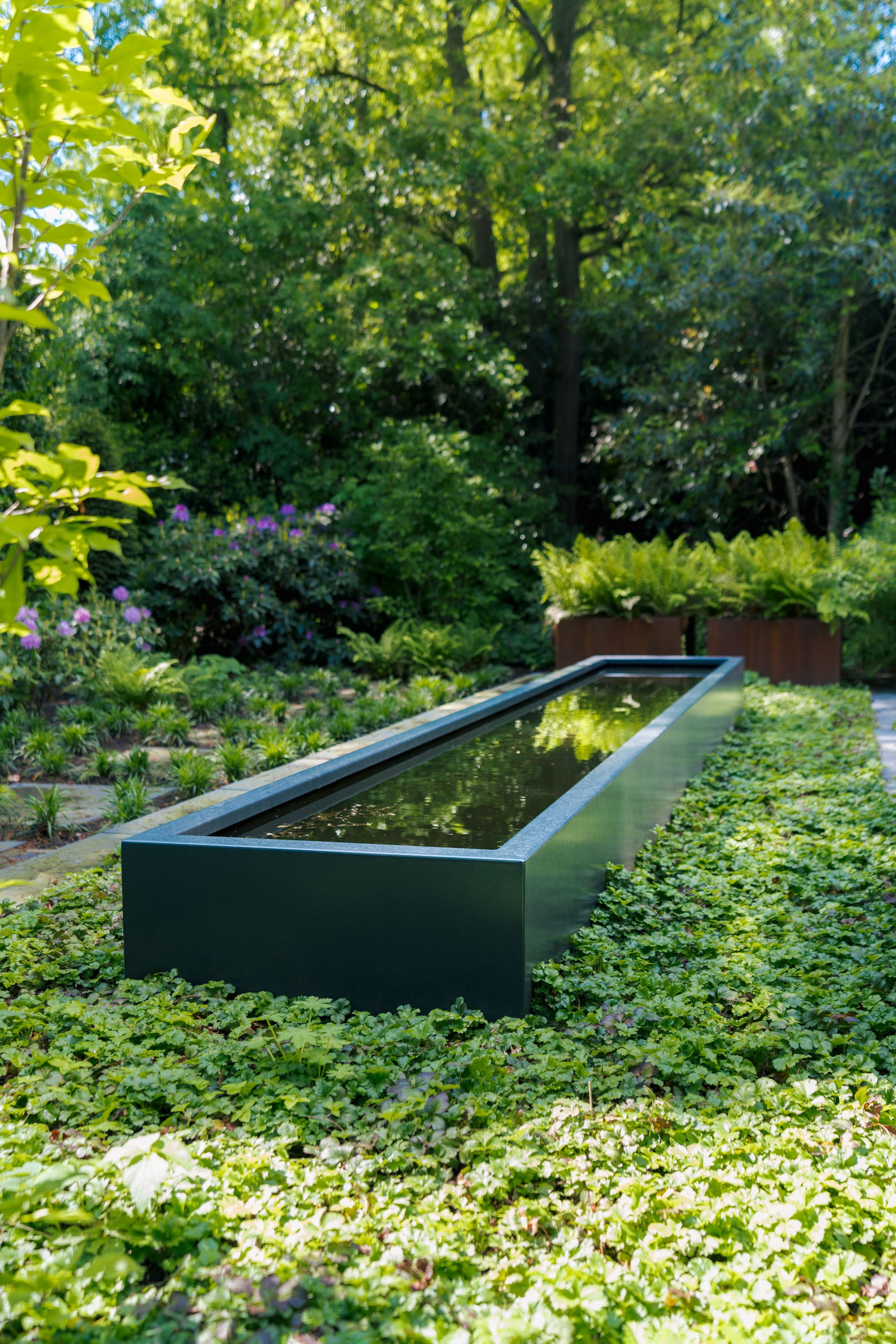 Long rectangular water feature in a garden with greenery and trees in the background